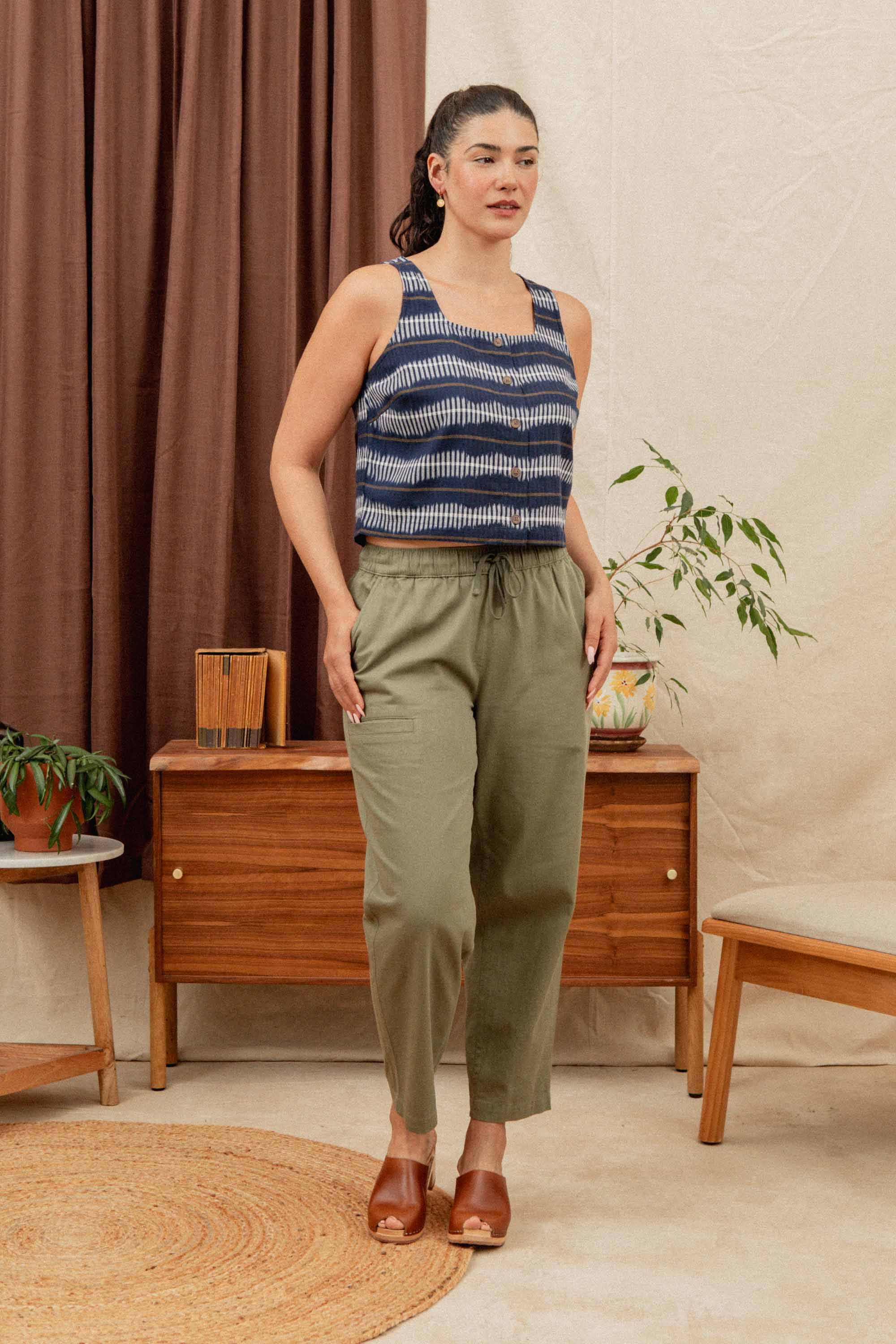 A woman stands indoors wearing the Bridge & Burn Sadie Tank in Dusk Mirage, paired with olive green pants and brown shoes, in a mid-century styled room with wooden furniture, plants, and neutral curtains.