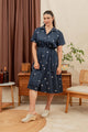A woman wears the Bridge & Burn Sylvie Dress in Lunar Polka—navy blue organic cotton with white circles—beside mid-century furniture, a plant, and brown curtains. Her hair is tied back and she pairs it with cream-colored shoes.