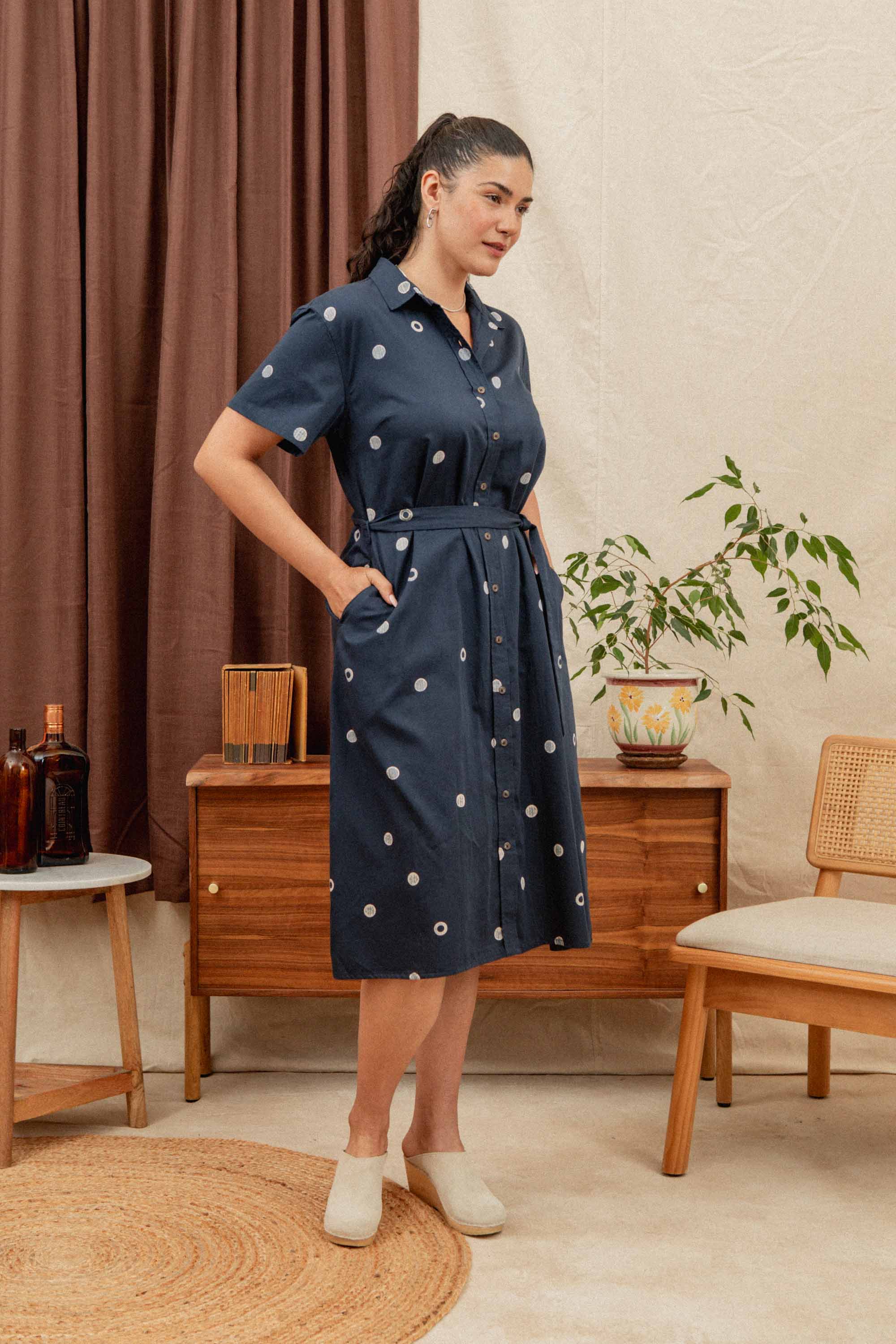 A woman wearing the Bridge & Burn Sylvie Dress in Lunar Polka stands in a mid-century modern room with wooden furniture, a potted plant, and beige decor. Her hands are in her pockets as she looks slightly to the side.
