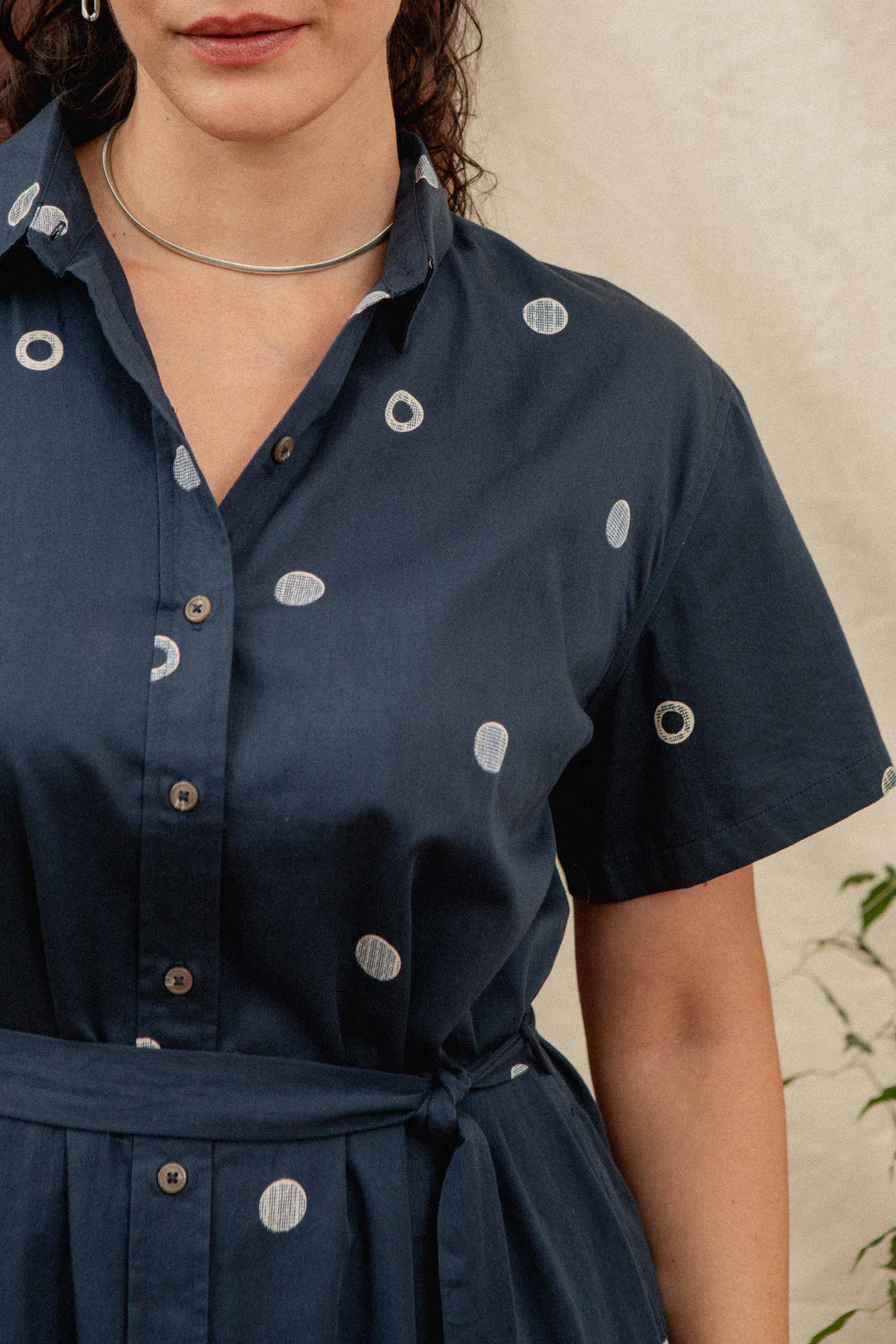 A woman wearing the Bridge & Burn Sylvie Dress in Lunar Polka—a navy blue, relaxed fit dress with white circles, button front, and a thin waist belt—stands partially in frame, accessorized with a silver choker and curly dark hair.