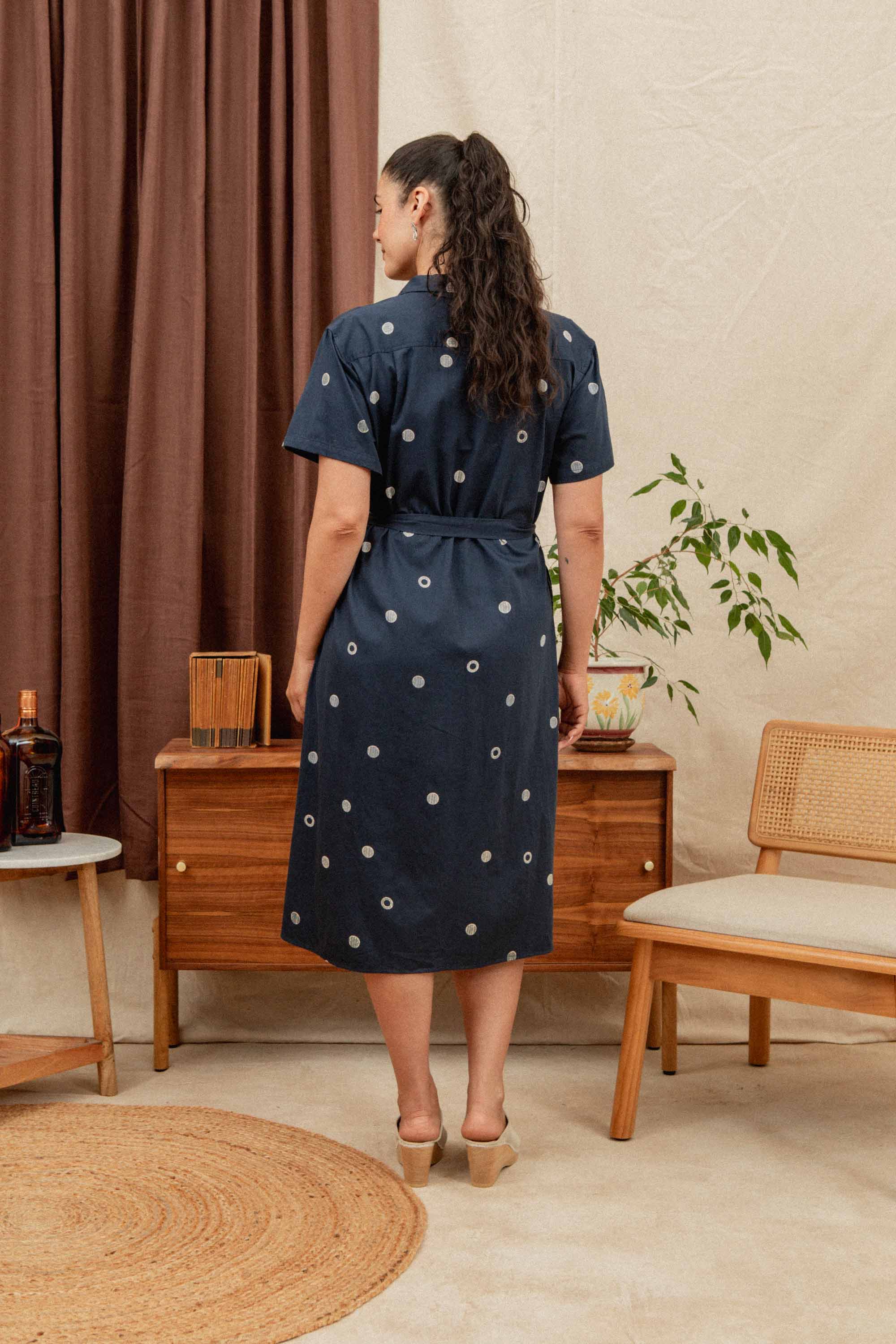 A woman with long dark hair in a ponytail stands with her back to the camera, wearing Bridge & Burn’s Sylvie Dress in Lunar Polka—a navy knee-length organic cotton shirtdress—in a mid-century modern room with wooden furniture and plants.