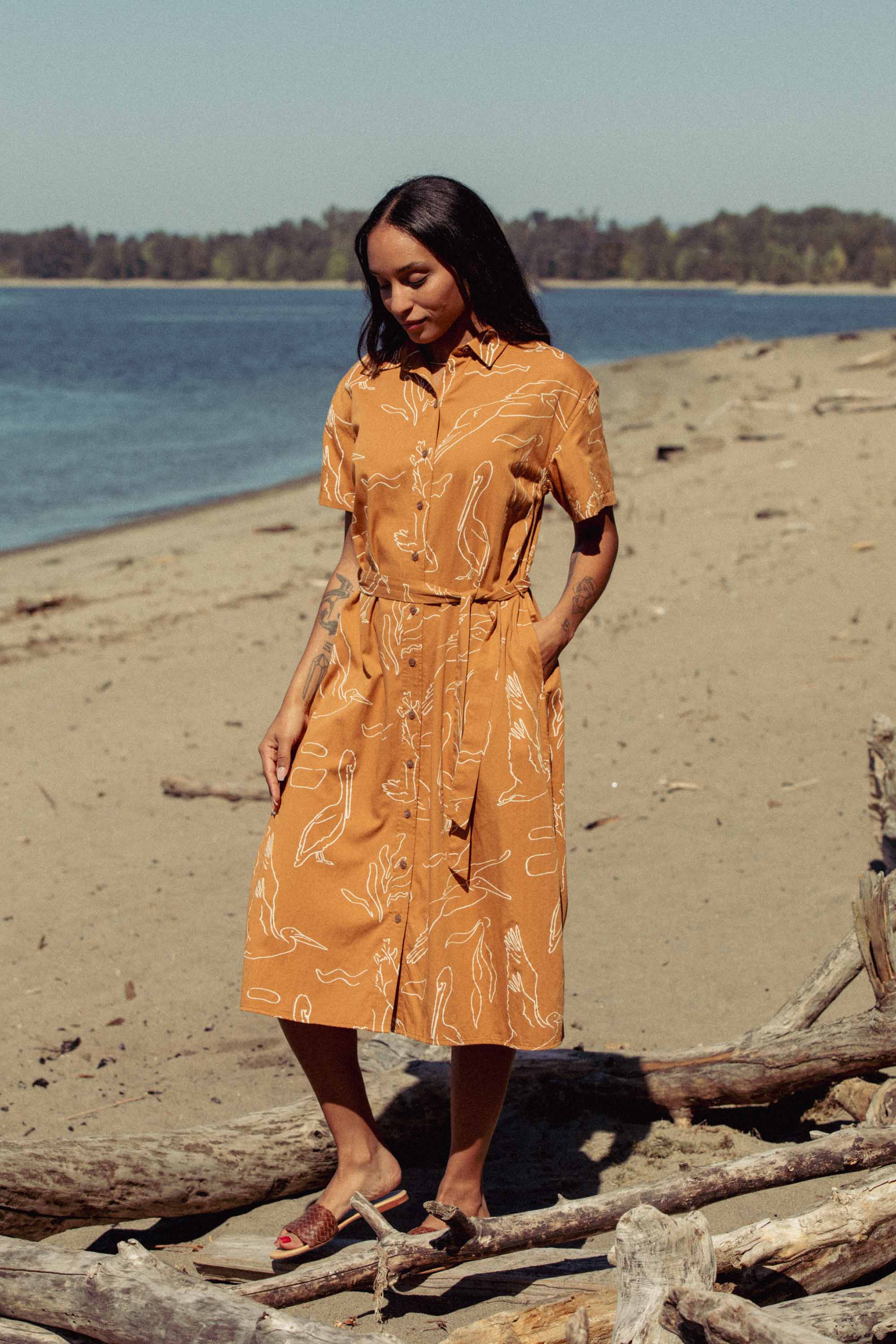 A woman wearing the Bridge & Burn Sylvie Dress in Shoreflight stands on a sandy beach near driftwood, with trees and water in the background under a clear sky.