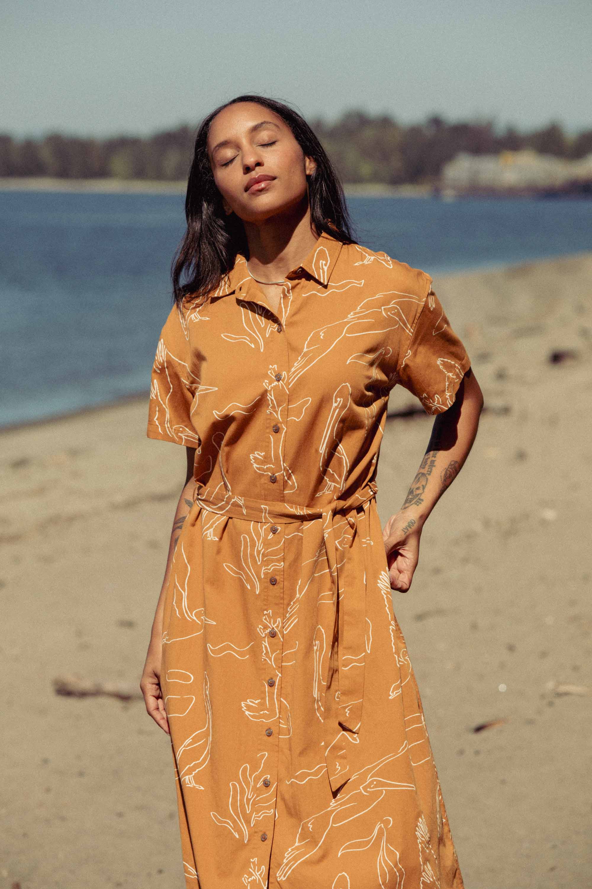 A woman stands on a sandy beach in the Bridge & Burn Sylvie Dress / Shoreflight, a mustard organic cotton dress with white abstract patterns. With closed eyes, she basks in the sunlight; trees and buildings appear in the background.