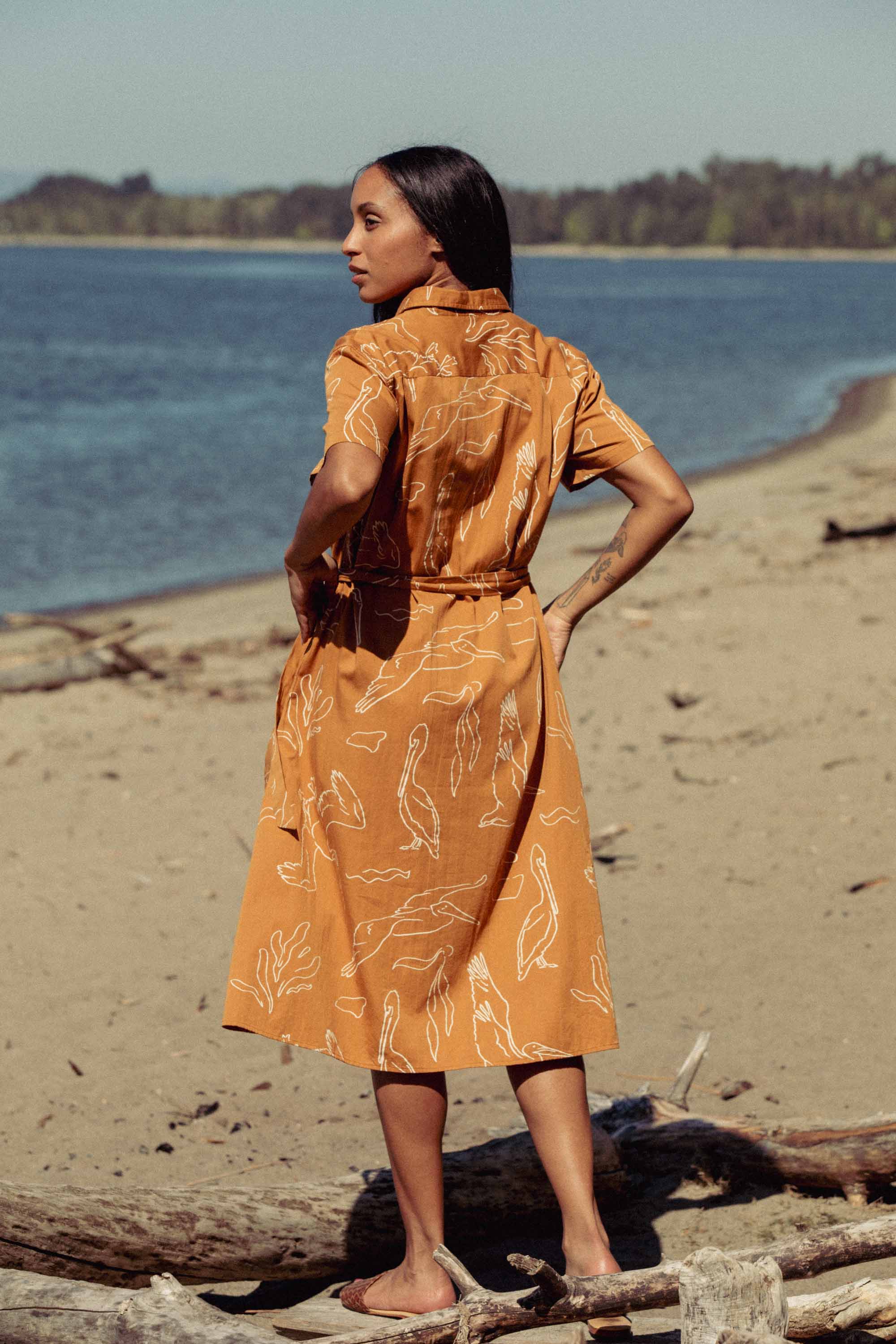 A woman stands barefoot on a sandy beach, facing away while wearing the Bridge & Burn Sylvie Dress in Shoreflight—a relaxed fit orange dress with white abstract lines—against a serene backdrop of water and distant trees.