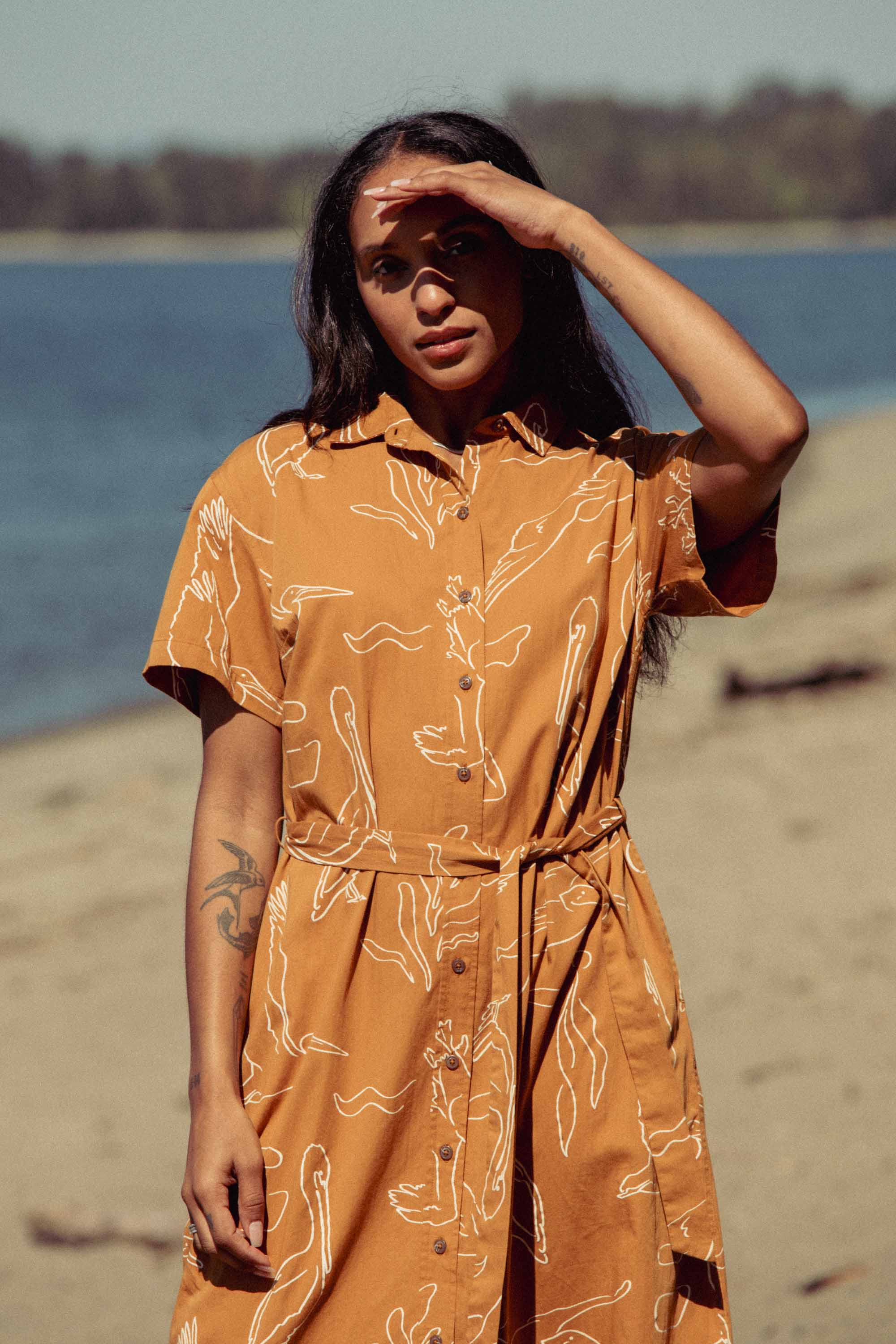 A woman in the Bridge & Burn Sylvie Dress in Shoreflight, an organic cotton orange patterned piece, stands on a sandy beach, shielding her eyes from the sun. Trees and water are visible in the background.