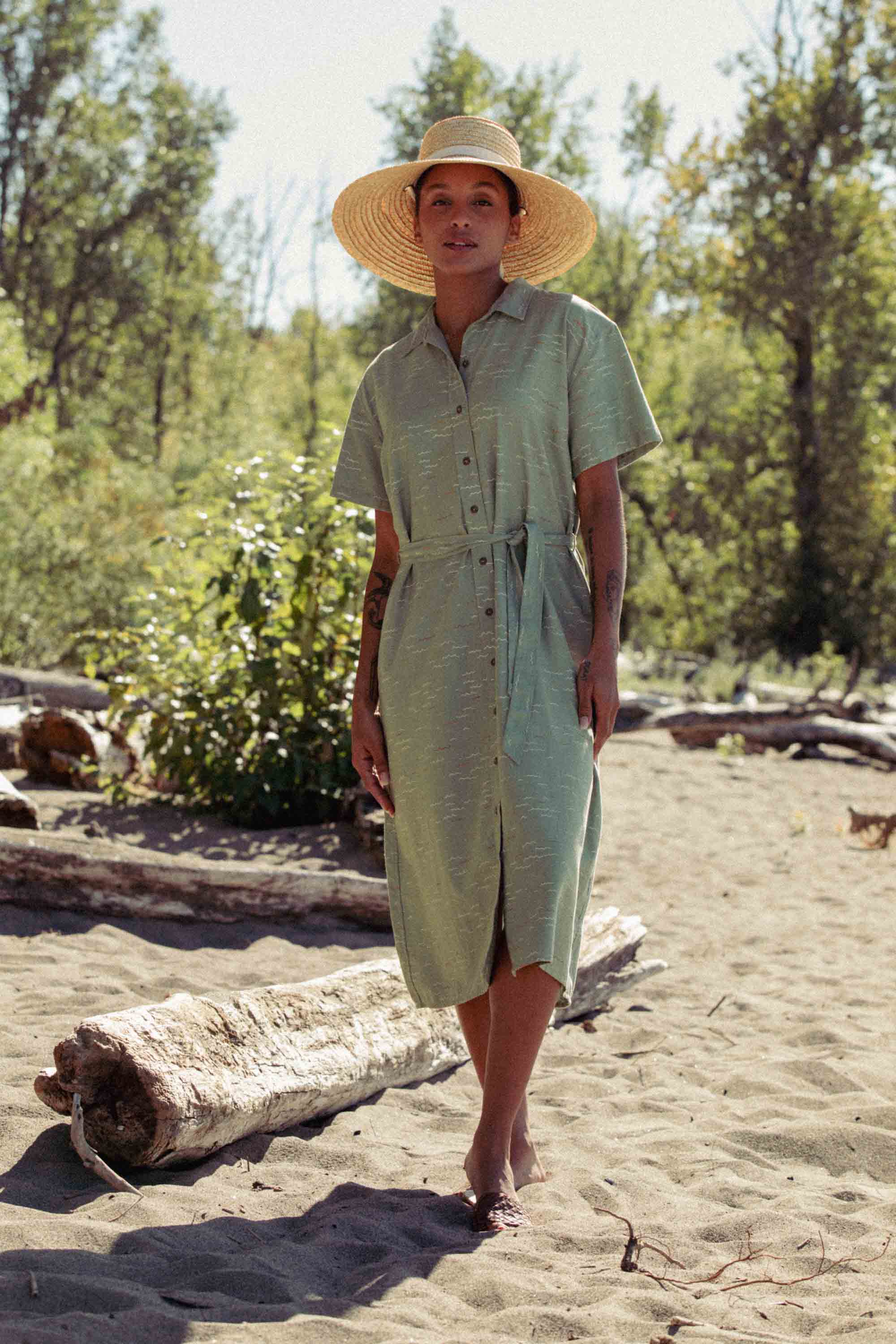Wearing the Bridge & Burn Sylvie Dress in Teal Tide Waves and a wide-brimmed straw hat, a person stands barefoot on sandy beach near driftwood, surrounded by trees and greenery under a sunny sky.