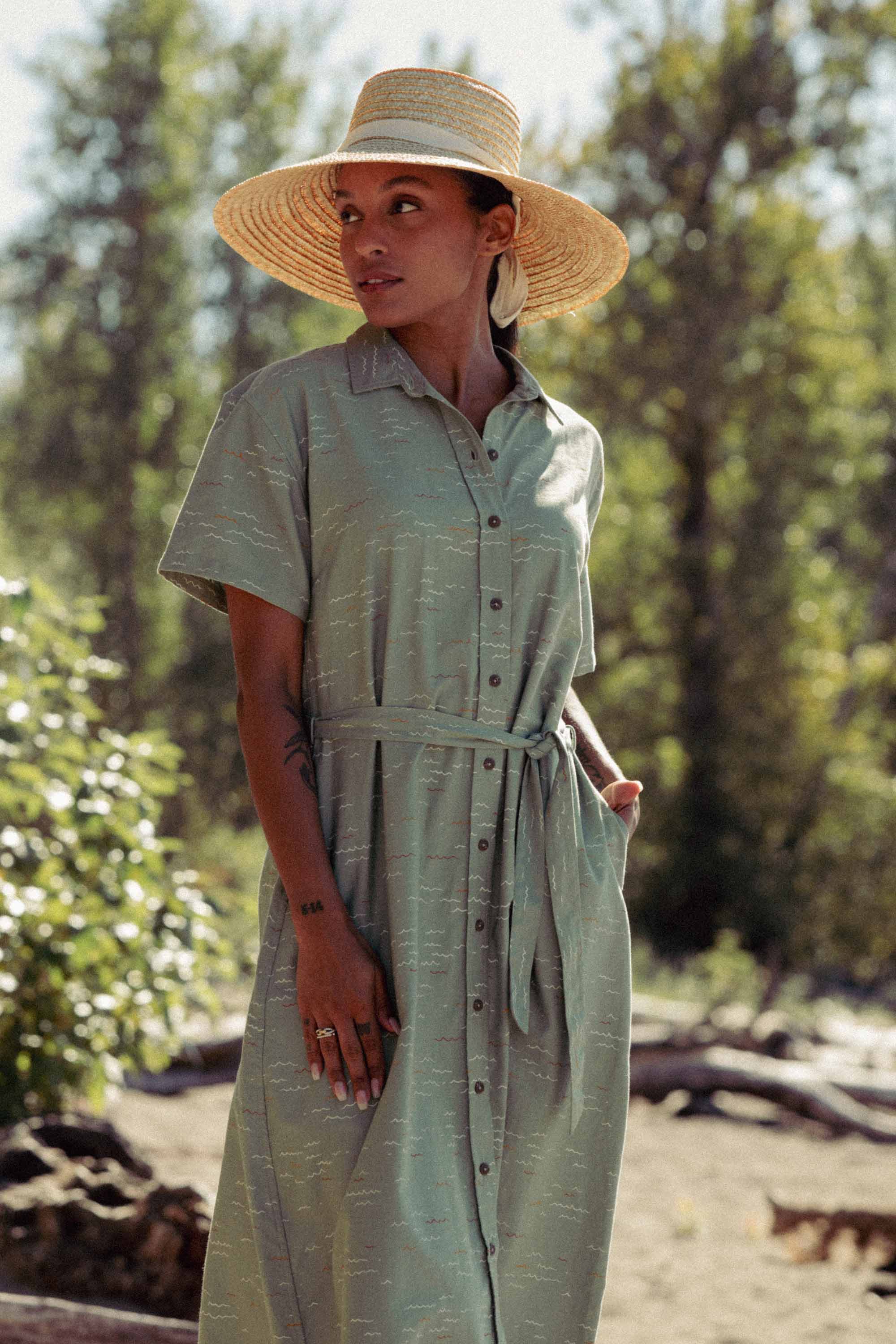 Wearing the Bridge & Burn Sylvie Dress in Teal Tide Waves, a person stands outdoors amid greenery and sunlight, accessorized with a wide-brimmed straw hat and featuring a relaxed fit with a waist tie.