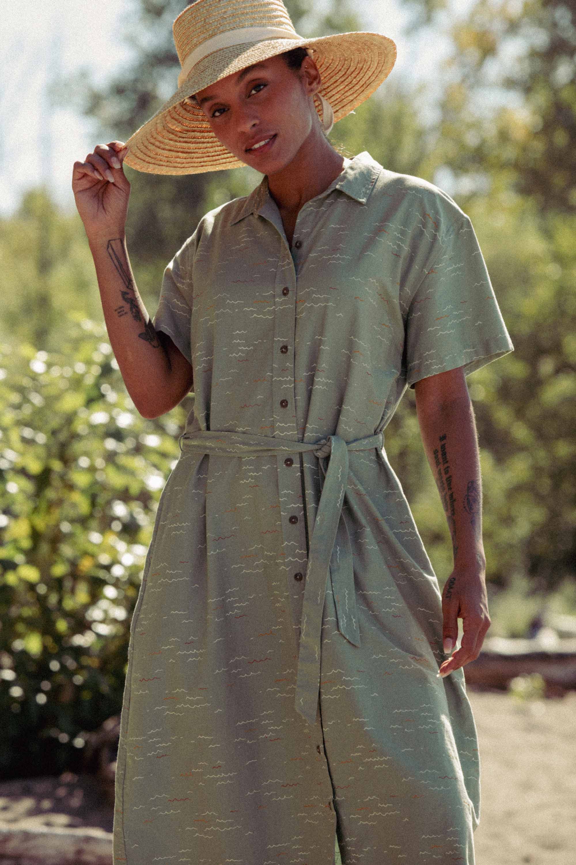 Wearing the Bridge & Burn Sylvie Dress in Teal Tide Waves, a person stands outdoors amid greenery and sunlight, holding the brim of a wide-brimmed straw hat with one hand.