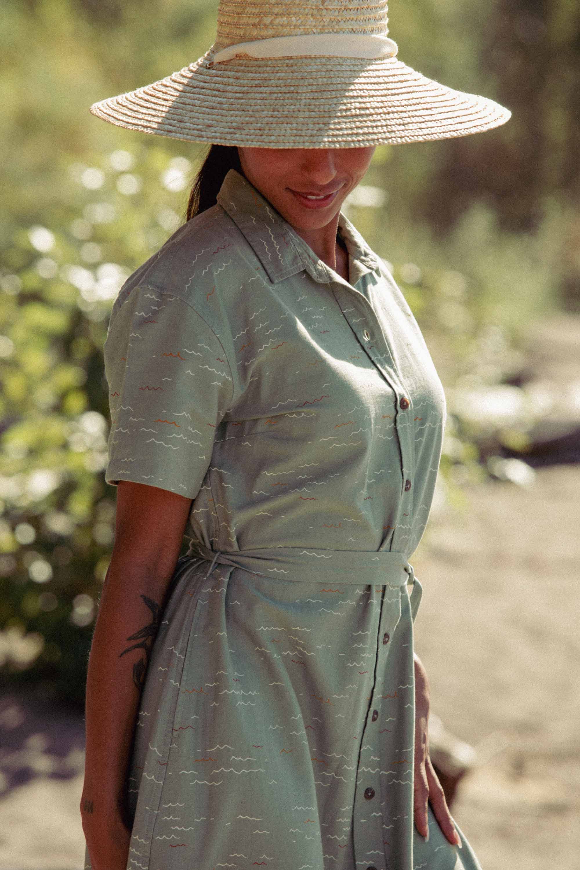 A woman in the Bridge & Burn Sylvie Dress in Teal Tide Waves stands outdoors with a wide-brimmed straw hat shading her face. A tattoo is visible on her left arm.