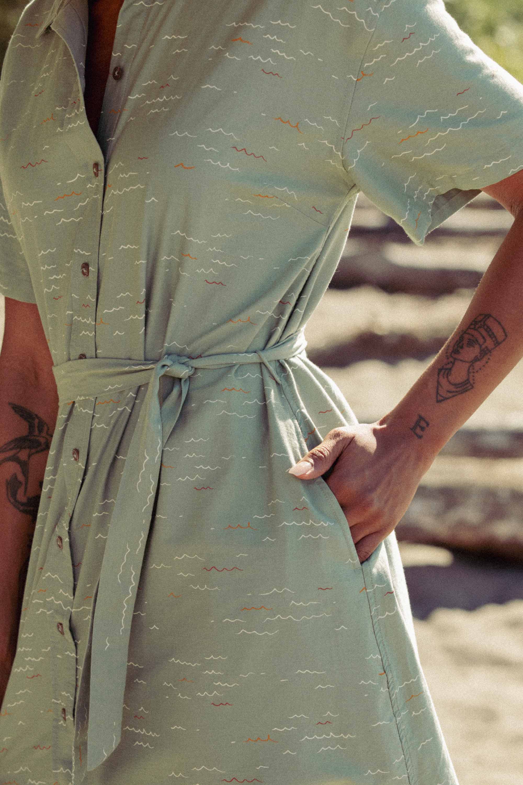 Outdoors, a person wears the Bridge & Burn Sylvie Dress in Teal Tide Waves—a light green organic cotton shirtdress with short sleeves, waist tie, pockets, and subtle wavy patterns. Forearm tattoos show as their hand rests in a pocket; logs blur behind.