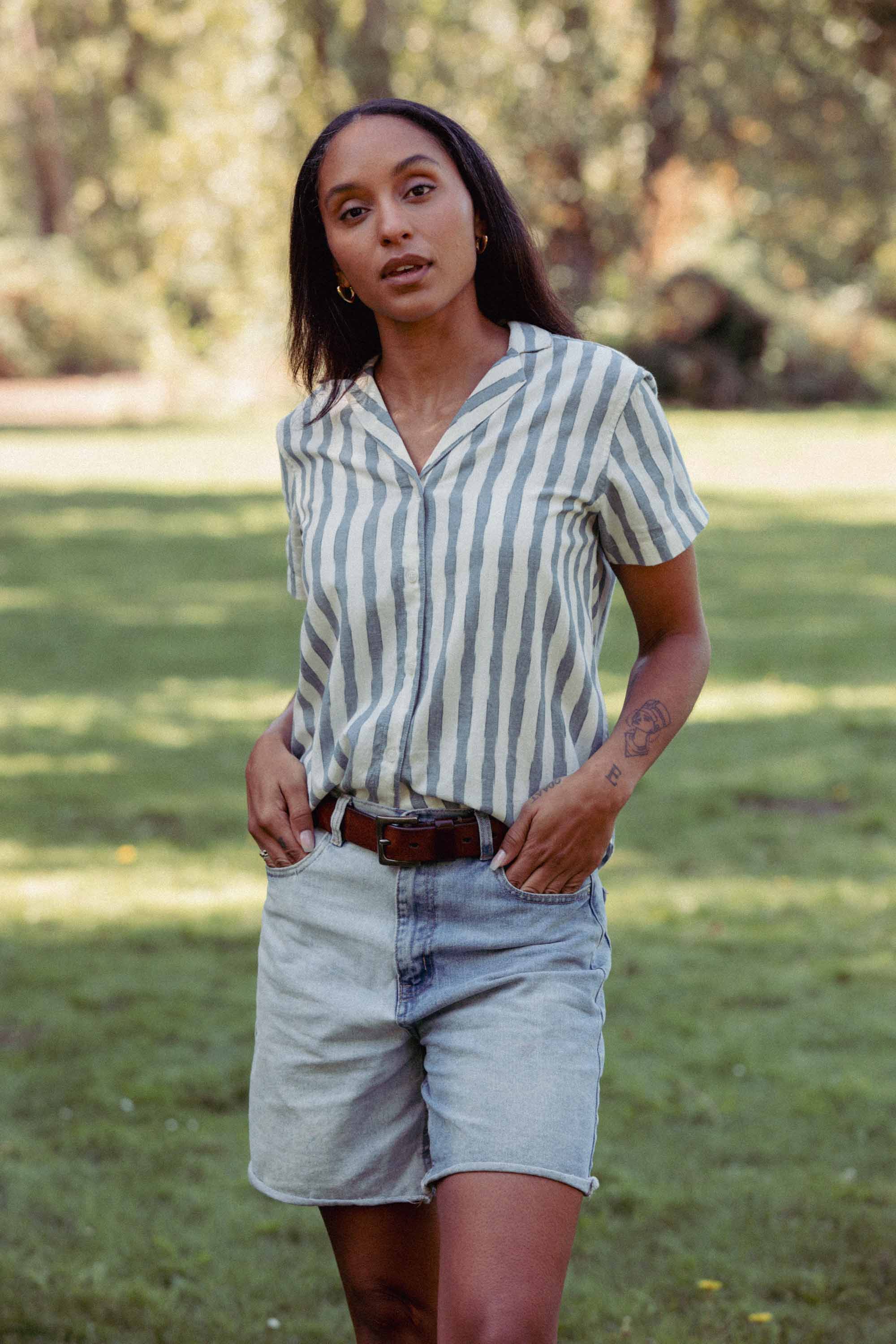 A woman stands on grass in sunlight, wearing the Bridge & Burn Wren Shirt in Beach Stripe with light denim shorts. She faces the camera with hands in her pockets, surrounded by trees.