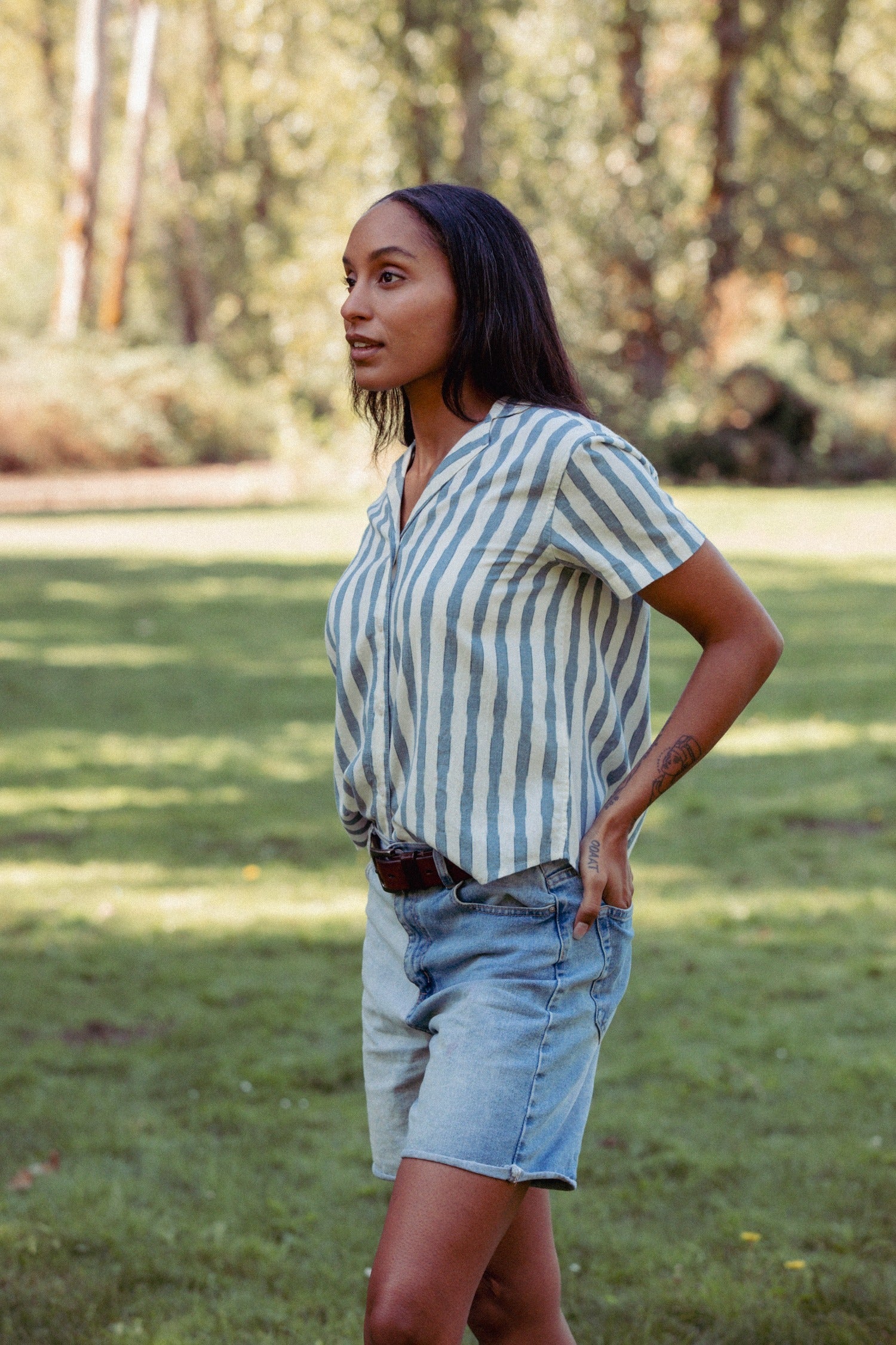 A woman stands on grass under the sun, wearing the Bridge & Burn Wren Shirt in Beach Stripe—white with blue stripes—and denim shorts. She has long dark hair and looks to the side, with trees in the background.