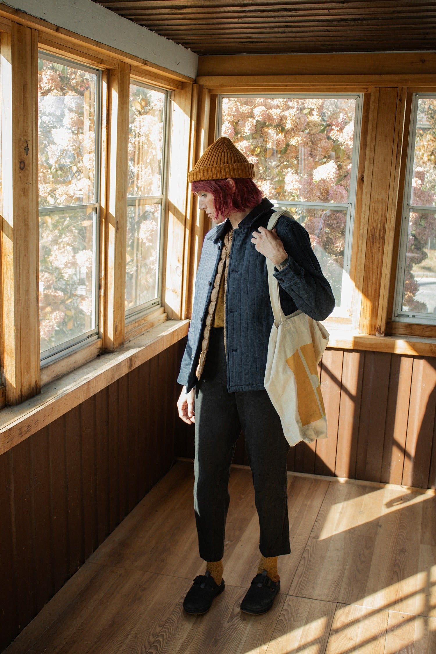 A person with red hair, wearing a yellow beanie and the Bridge & Burn Kenton Jacket in Slate with dark pants, stands in a sunlit wooden room holding a cream tote bag and gazing out large windows.