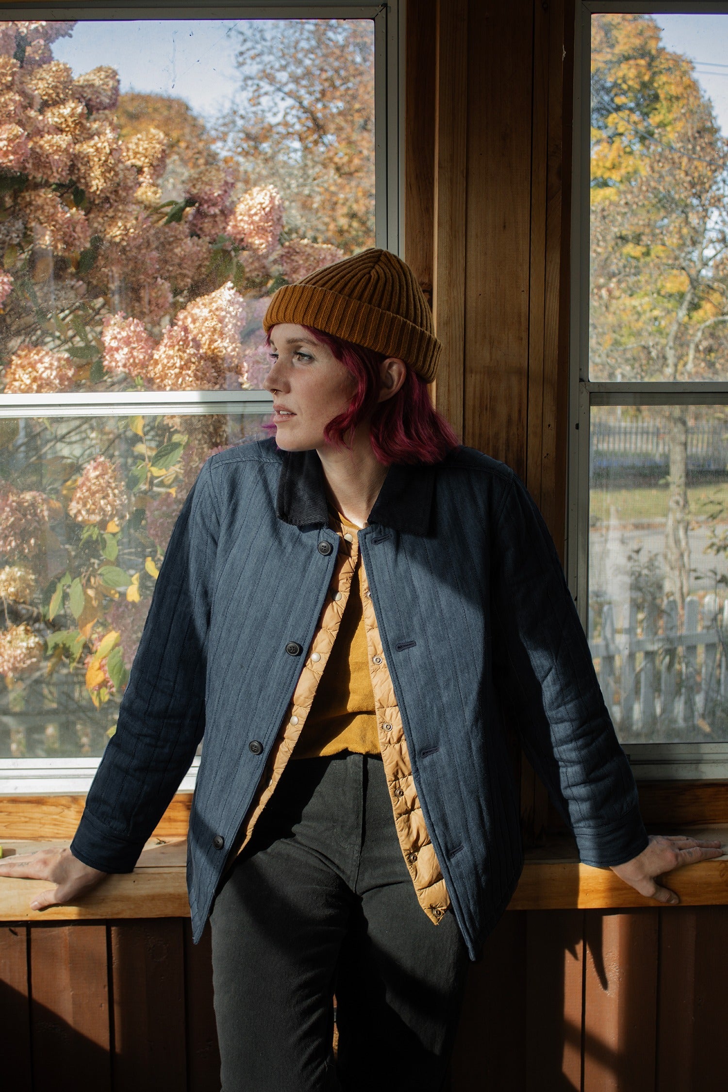 A person with pink hair, wearing a mustard beanie and shirt, leans against a sunlit windowsill in a blue Kenton Jacket by Bridge & Burn. Autumn trees with pink leaves can be seen outside.