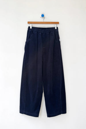Le Bon Shoppe's Arc Pant in Midnight Corduroy features a wide-leg, high-waist design with an elastic waistband and cropped legs, shown on a wooden hanger against a plain white background.
