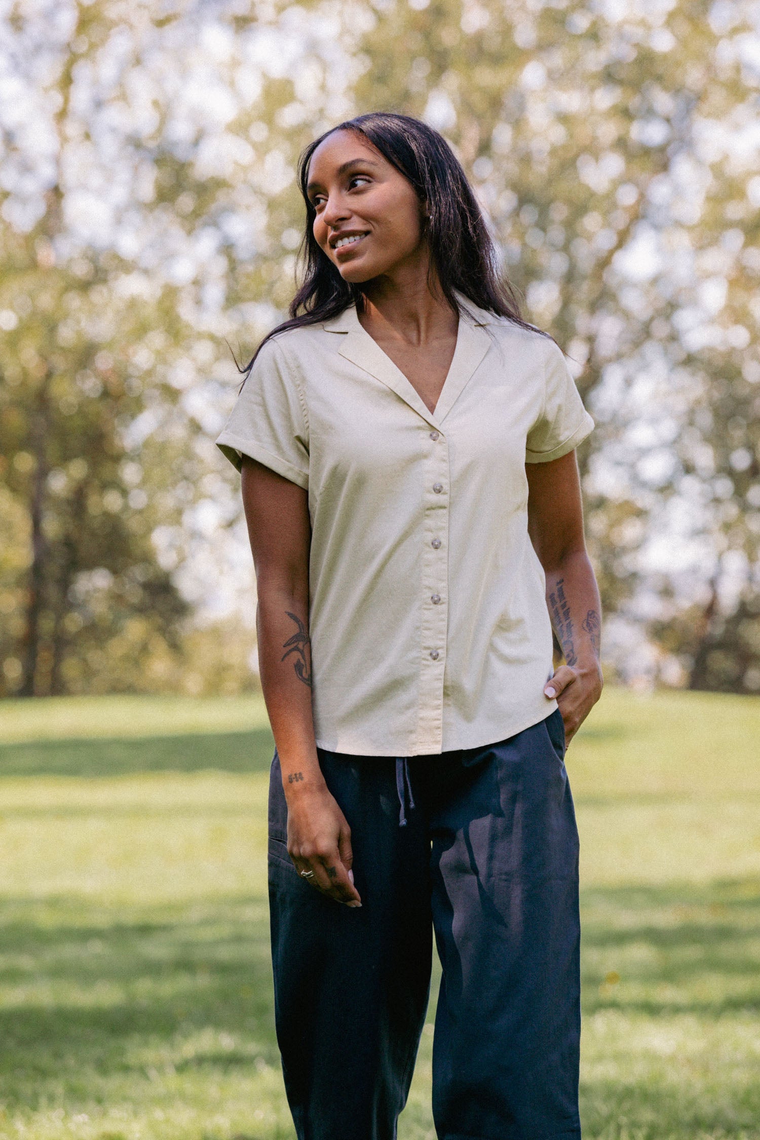 A woman with long dark hair stands outdoors on grass, smiling to the side and wearing the Bridge & Burn Innes Shirt in Ivory with dark pants; trees and sunlight provide a warm, natural backdrop.