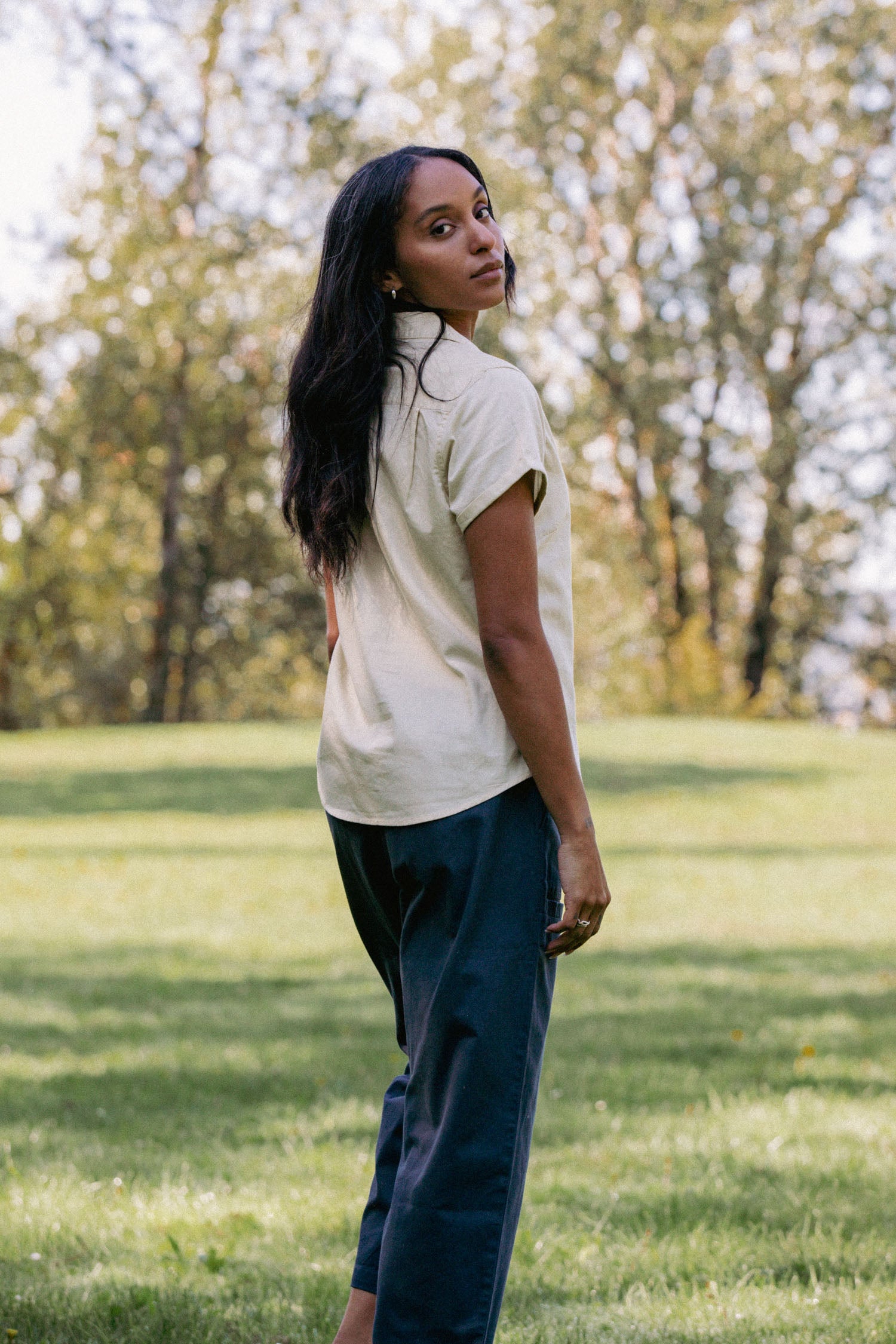 A woman with long dark hair stands on grass outdoors, wearing the Bridge & Burn Innes Shirt in Ivory and dark pants, looking back over her shoulder. Soft-focus green trees fill the background.