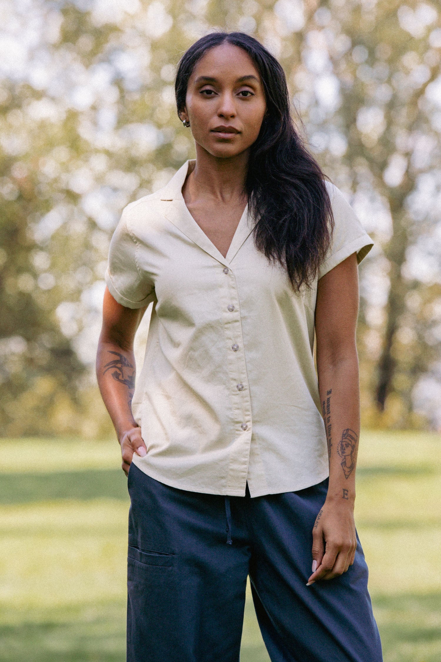 Outdoors, a person with long dark hair and visible arm tattoos stands with hands in pockets, wearing the Bridge & Burn Innes Shirt in Ivory and dark pants. Trees and greenery create a blurred background.