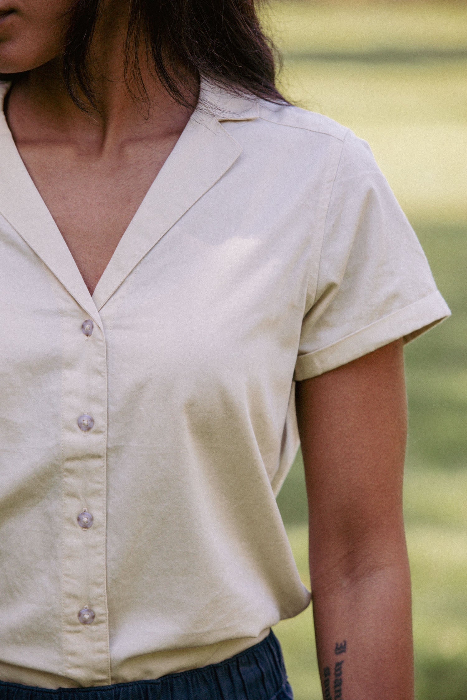 A person stands on grass wearing the Bridge & Burn Innes Shirt in Ivory—a short sleeve, relaxed fit button-up blouse. The image shows their upper body and right arm, featuring a visible tattoo.