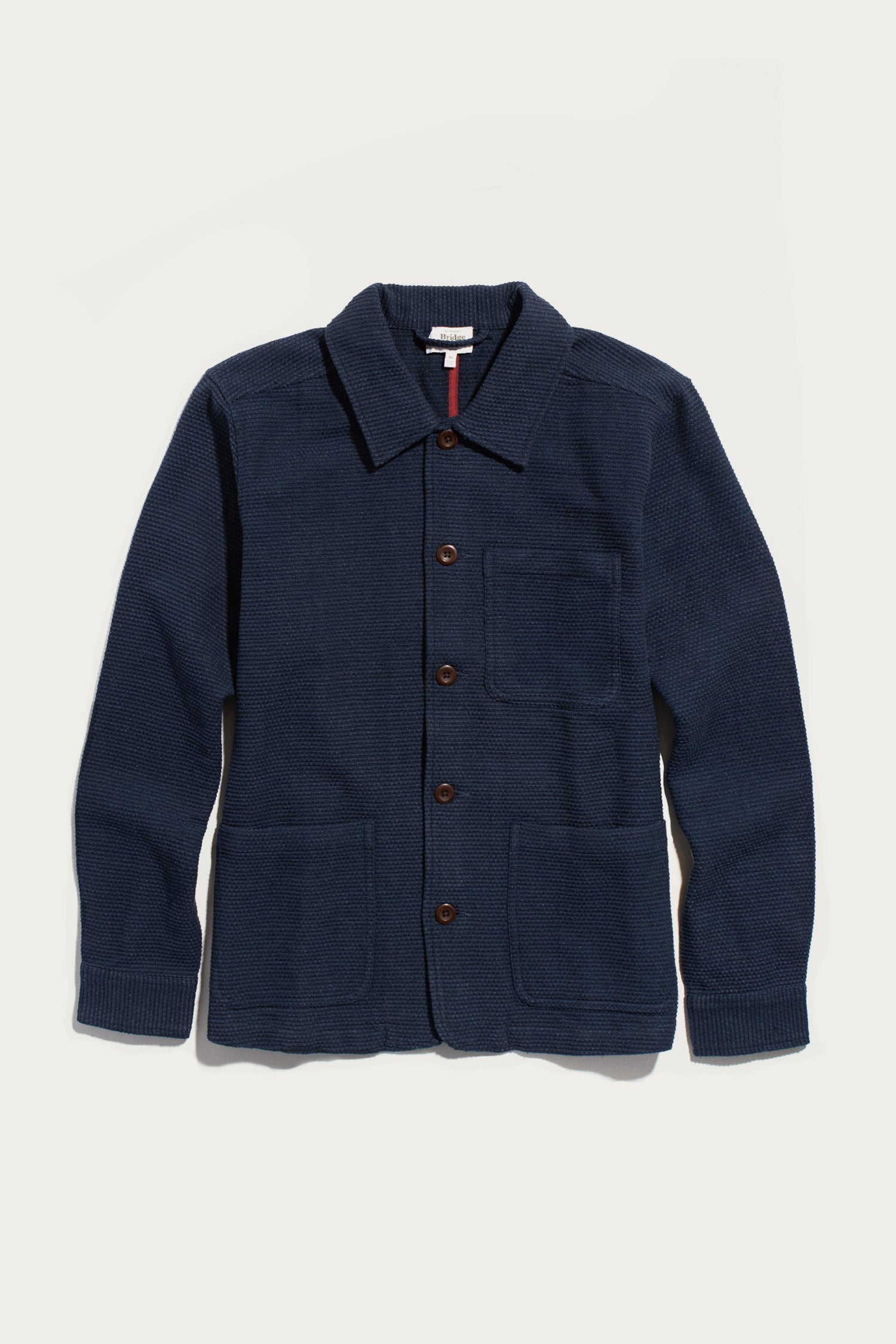 The Bridge & Burn Men’s Studio Chore Coat in Indigo is inspired by classic chore coat design, featuring a collar, chest pocket, and two large front pockets. Crafted from organic cotton, it is displayed on a white background.