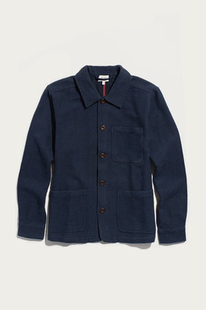 The Bridge & Burn Men’s Studio Chore Coat in Indigo is inspired by classic chore coat design, featuring a collar, chest pocket, and two large front pockets. Crafted from organic cotton, it is displayed on a white background.