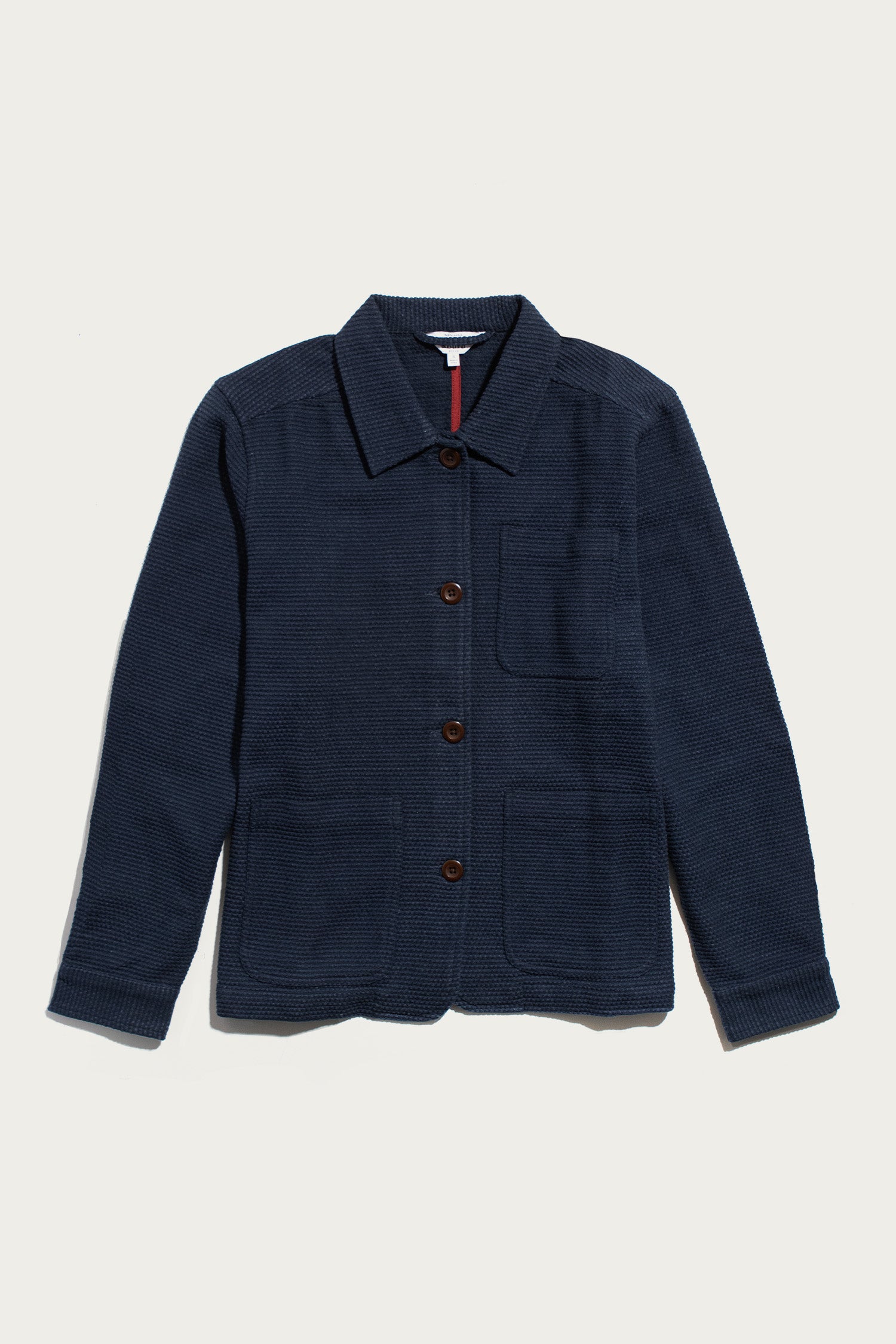 Bridge & Burn’s Women’s Studio Chore Coat in indigo is a navy blue textured workwear jacket featuring a collar, chest pocket, and two large front pockets, crafted from organic cotton and shown laid flat on a white background.