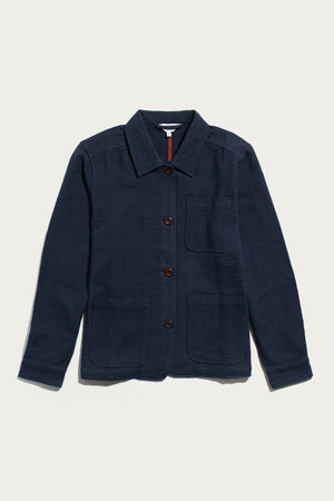 Bridge & Burn’s Women’s Studio Chore Coat in indigo is a navy blue textured workwear jacket featuring a collar, chest pocket, and two large front pockets, crafted from organic cotton and shown laid flat on a white background.
