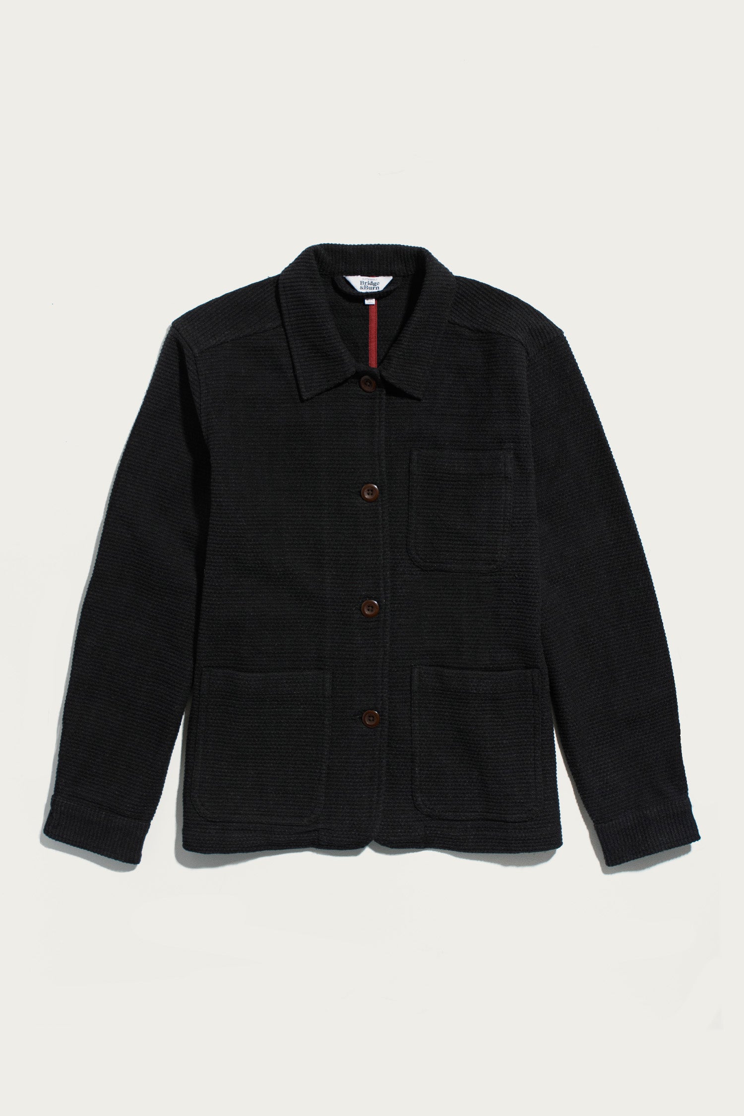 The Women’s Studio Chore Coat by Bridge & Burn in washed black—crafted from organic cotton—features a collar, three front pockets, and brown buttons, shown laid flat on a white background.