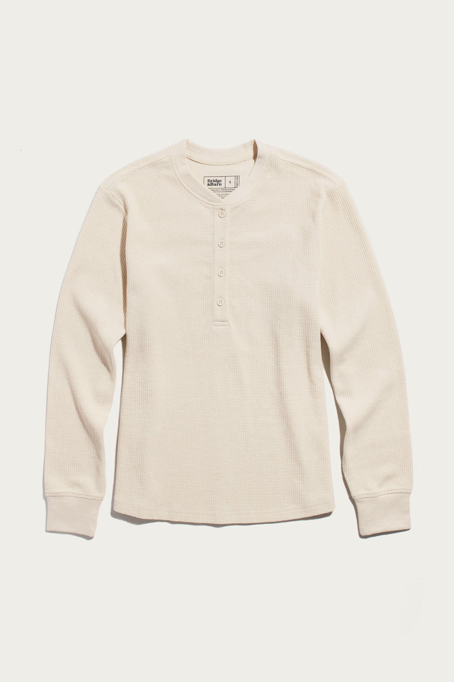 The Women’s Recycled Waffle Henley in Ivory by Bridge & Burn, a long-sleeve shirt made from recycled fibers with a crew neckline and four-button placket, is laid flat on a white background.