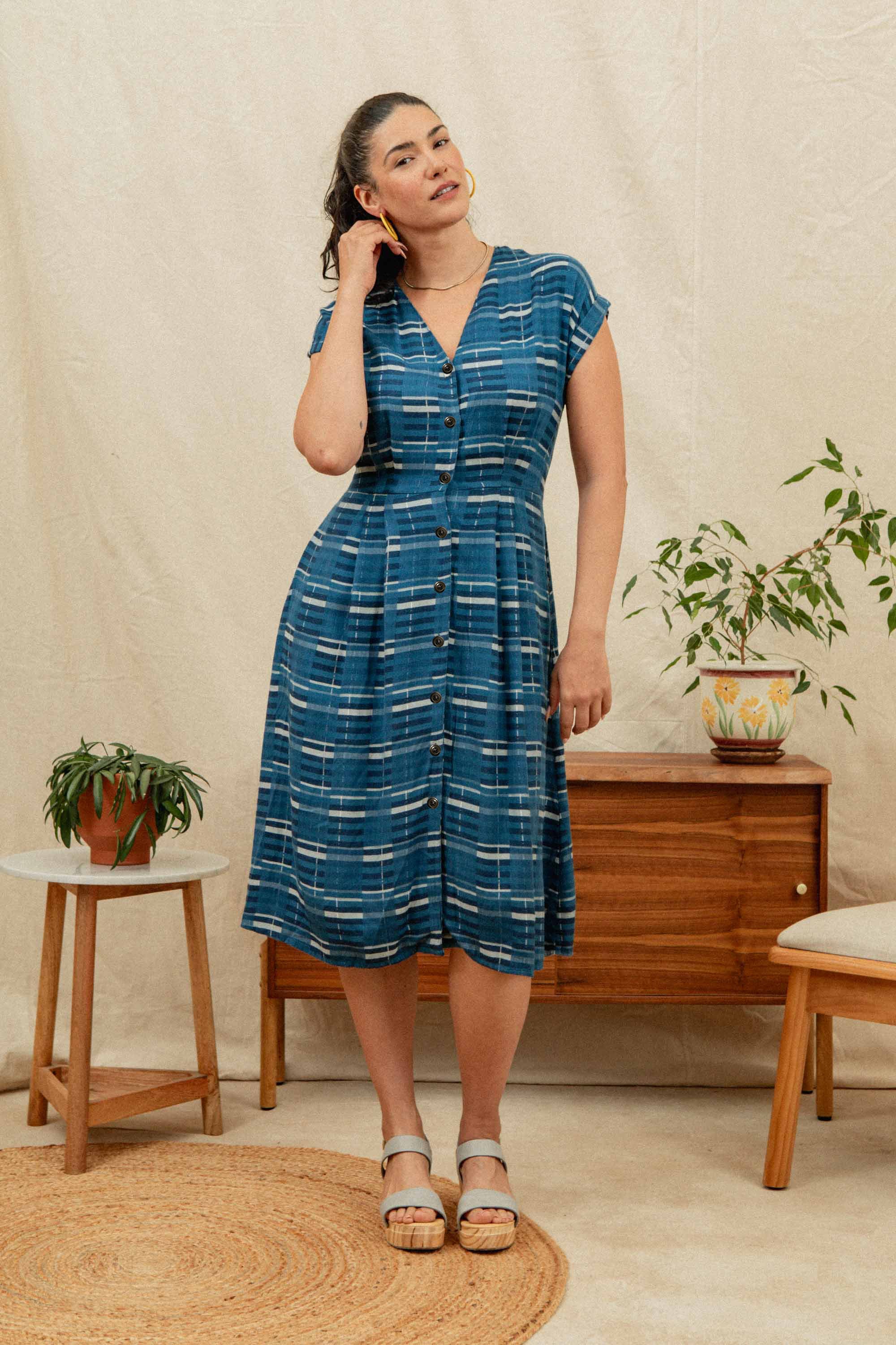 A woman wears the Bridge & Burn Eileen Dress in Stormcrest Stripe, standing indoors by a wooden dresser with potted plants, touching her earring. She stands on a round woven rug against a light-colored wall.
