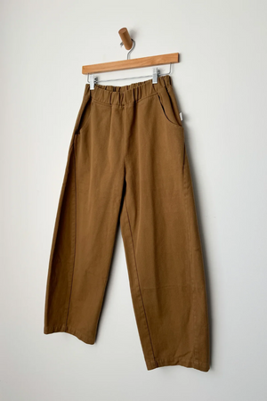The Le Bon Shoppe - Arc Pant in Tobacco features a wide-leg fit, heavy-weight canvas, elastic waistband, and side pockets, shown hanging on a wooden hanger against a plain white wall.