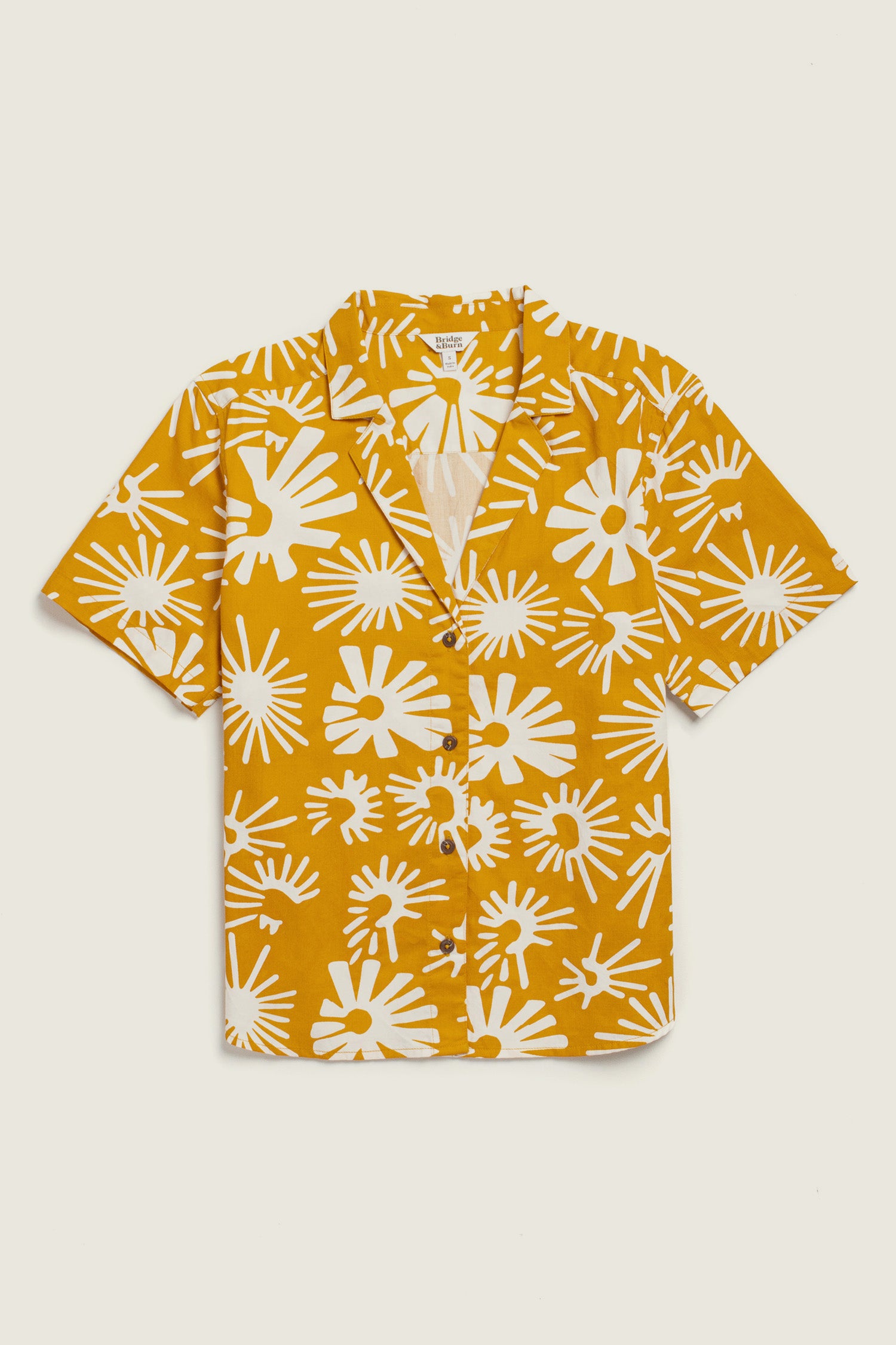 The Mila Cropped Shirt / Solar Bloom by Bridge & Burn is a short-sleeved, button-up mustard yellow shirt with a white abstract starburst print, crafted from organic cotton for a relaxed fit and effortless comfort.