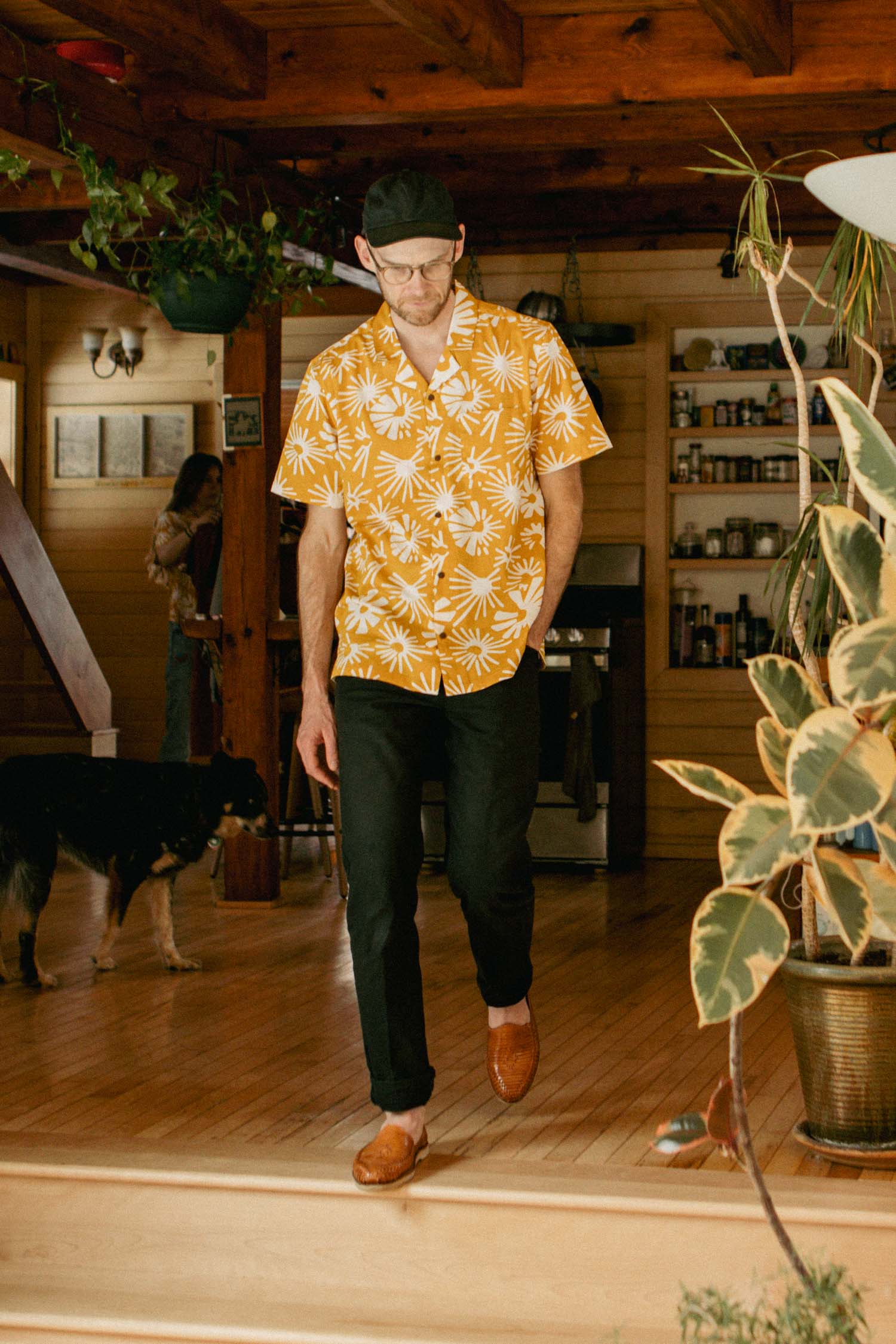 A man wears the Bridge & Burn Bodie Shirt in Solar Bloom with black pants and brown shoes, walking indoors on wooden flooring. A dog and another person are in the background among plants, wooden beams, and shelves with jars.