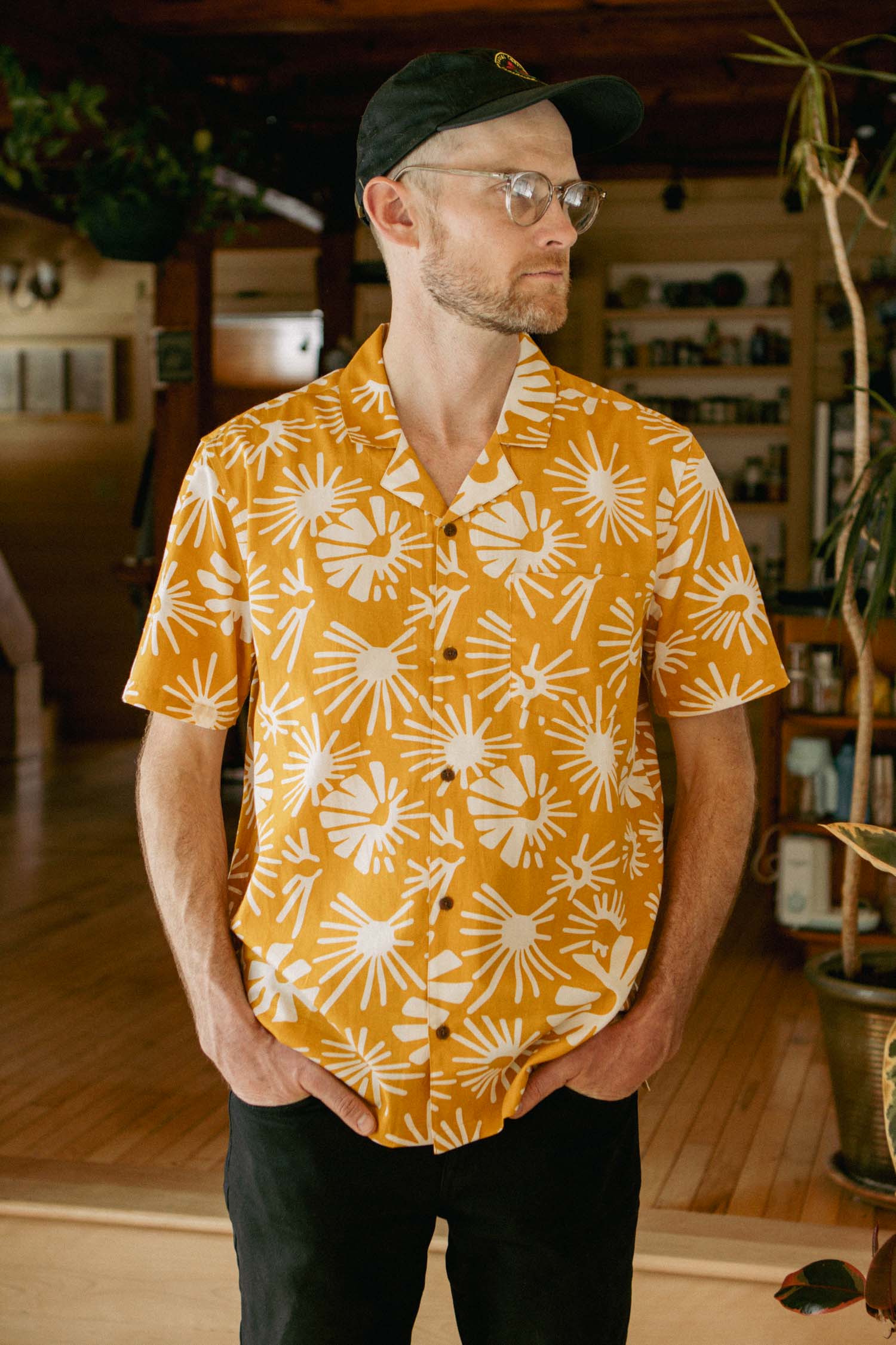 A man in glasses and a black cap stands indoors on wooden floors with plants, wearing the Bridge & Burn Bodie Shirt in Solar Bloom—a retro-inspired yellow camp collar shirt with a bold white pattern—and looks to the side with his hands in his pockets.