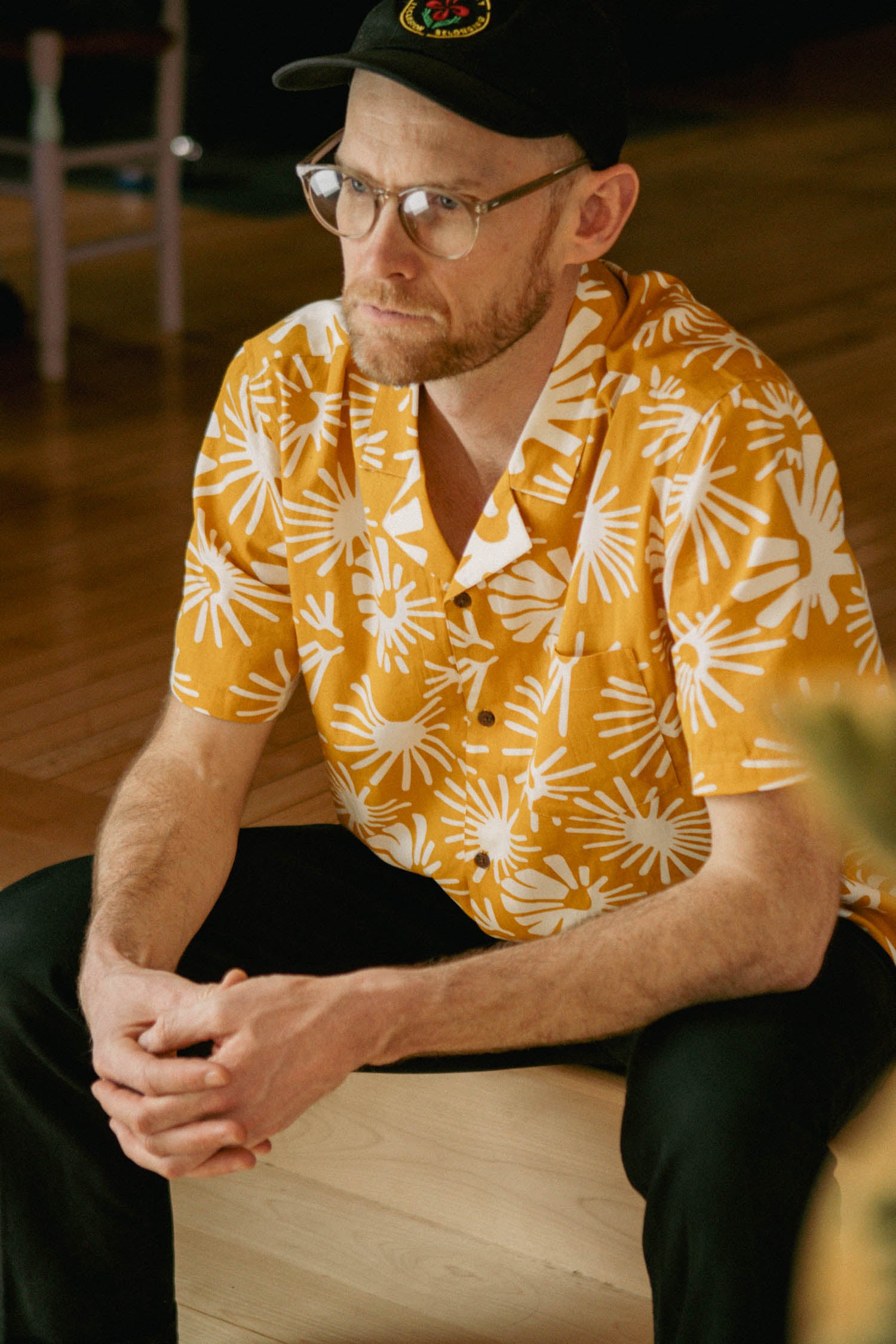 A man in glasses and a black cap sits indoors on a wooden floor, looking to the side with hands clasped. He wears the Bodie Shirt in Solar Bloom by Bridge & Burn—a yellow retro-inspired shirt with a white abstract pattern and camp collar.