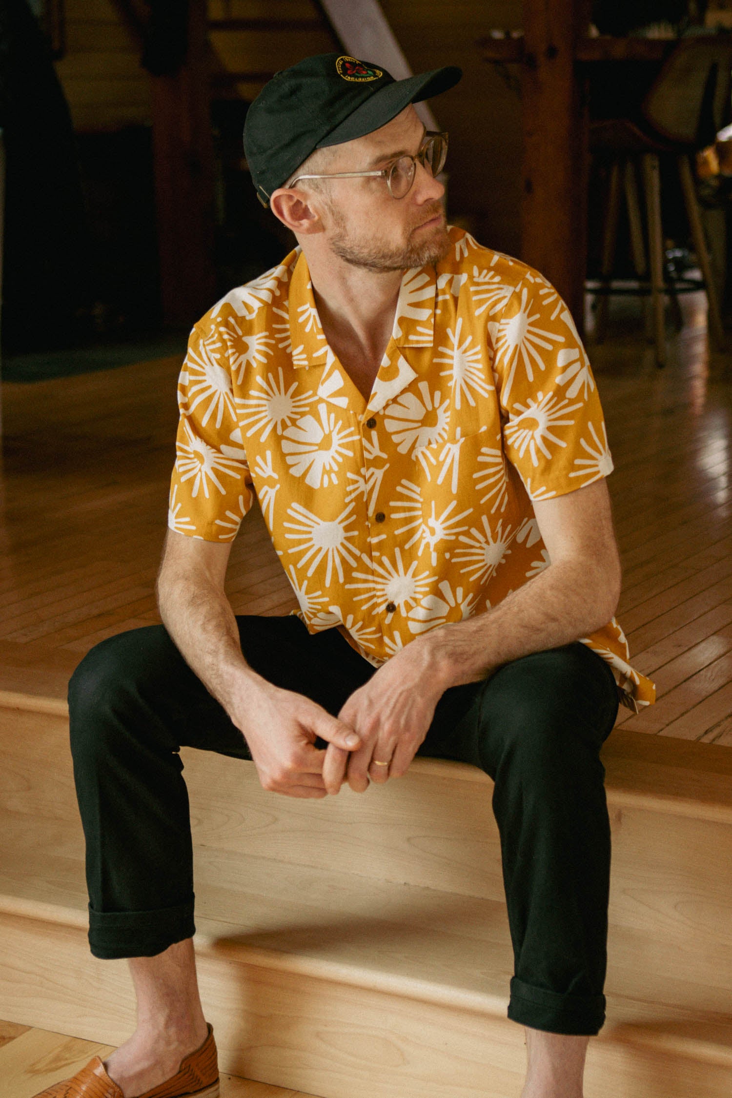 A man in the Bridge & Burn Bodie Shirt / Solar Bloom, black pants, glasses, and a black cap sits on wooden steps indoors, looking to his left.