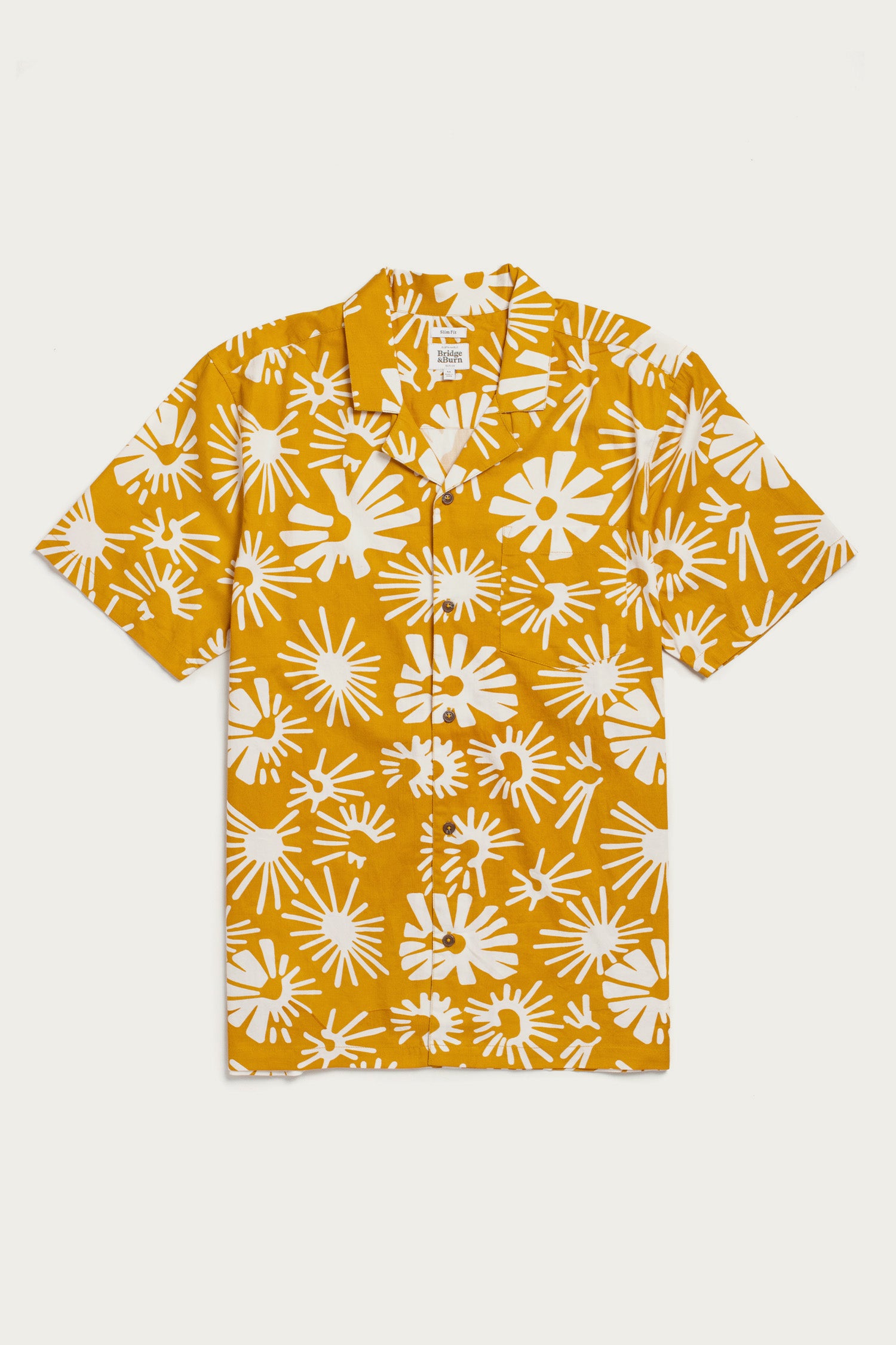 The Bodie Shirt / Solar Bloom by Bridge & Burn is a short-sleeved, organic cotton camp collar shirt featuring bold white abstract florals on a mustard yellow background, shown laid flat on a white surface.