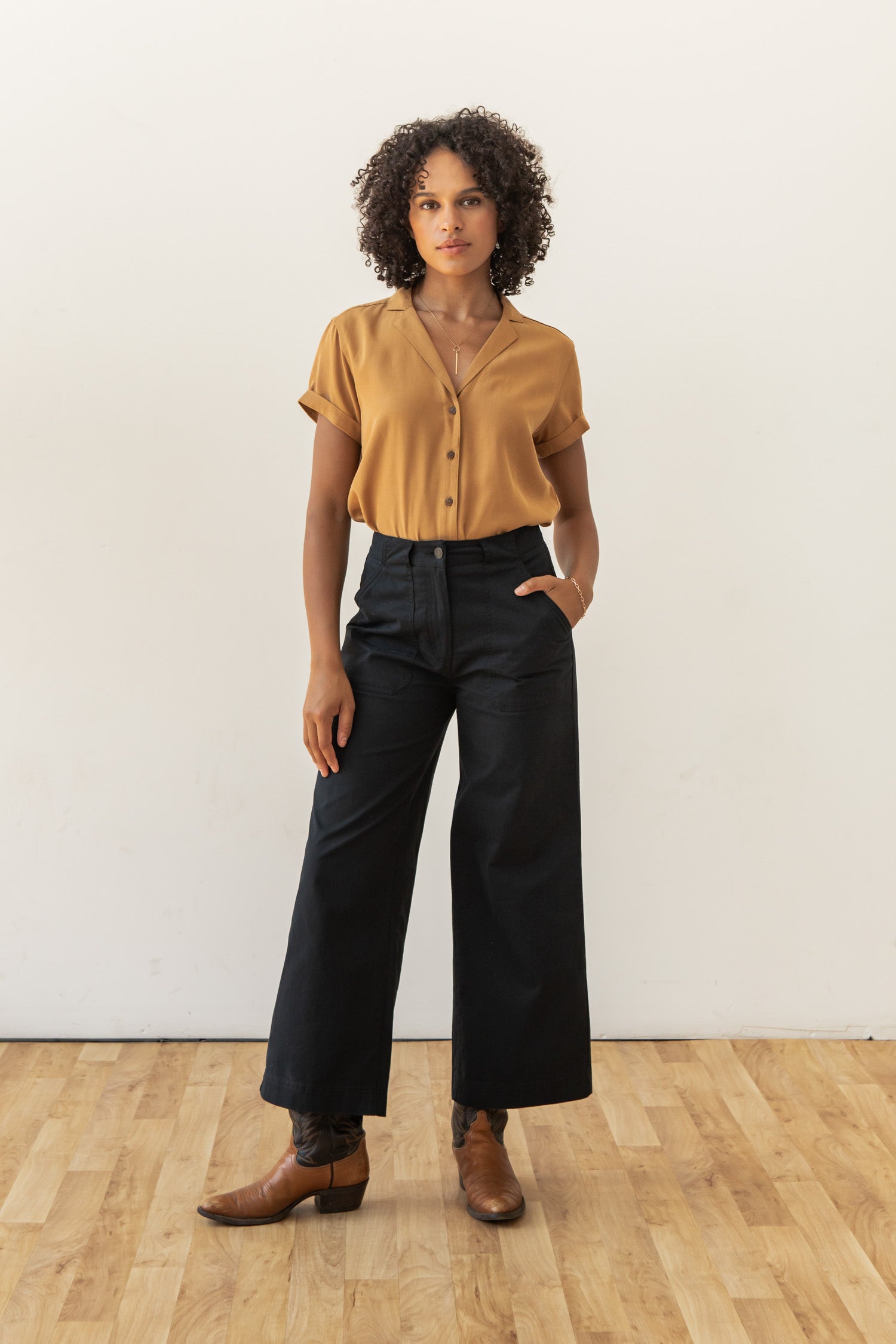 A person stands on a wooden floor against a white wall, wearing the Bridge & Burn Innes Shirt in Gold, wide-leg black pants, and brown ankle boots. One hand rests in their pocket as they look straight ahead.
