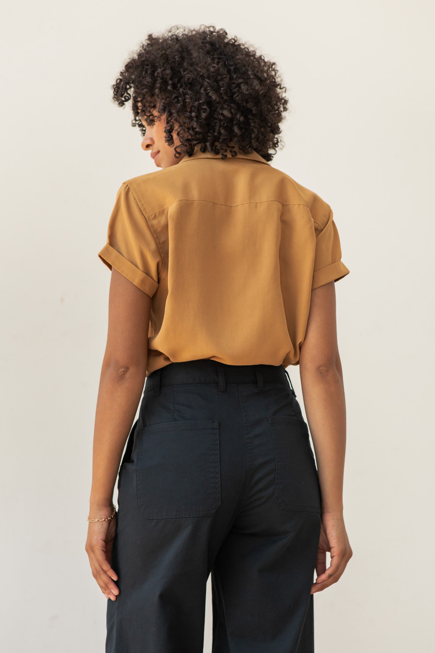 A person with curly hair, wearing the Bridge & Burn Innes Shirt in gold and high-waisted black pants, stands facing away from the camera against a plain light background.