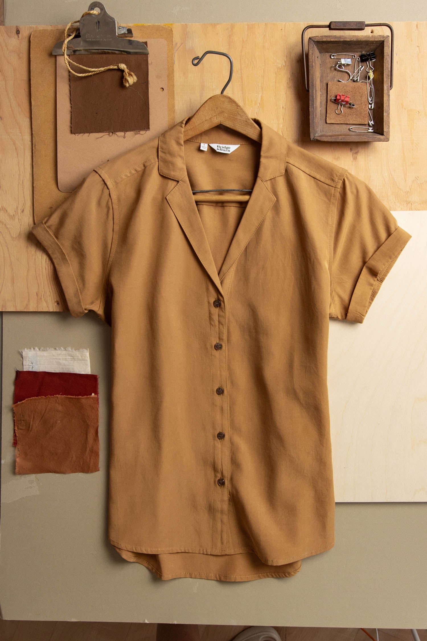 The Bridge & Burn Innes Shirt in Gold, an organic cotton short-sleeve button-up blouse, hangs on a wooden background surrounded by fabric swatches, thread, and sewing tools.