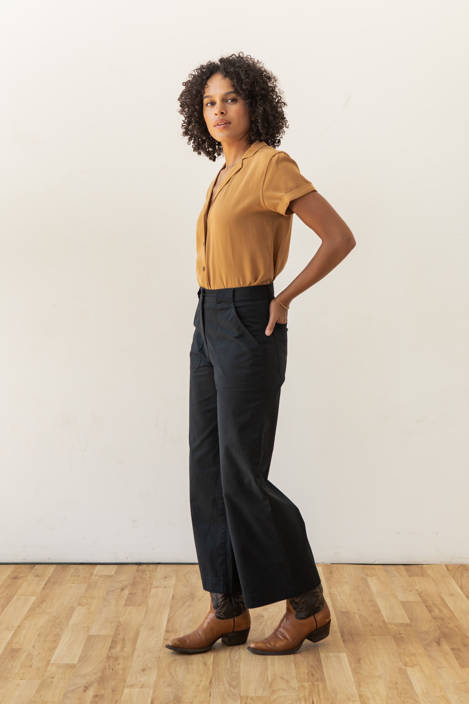 A woman with curly hair stands confidently indoors on a wooden floor, wearing the Bridge & Burn Innes Shirt in gold with high-waisted dark pants and brown pointed-toe shoes, posing before a plain white wall.