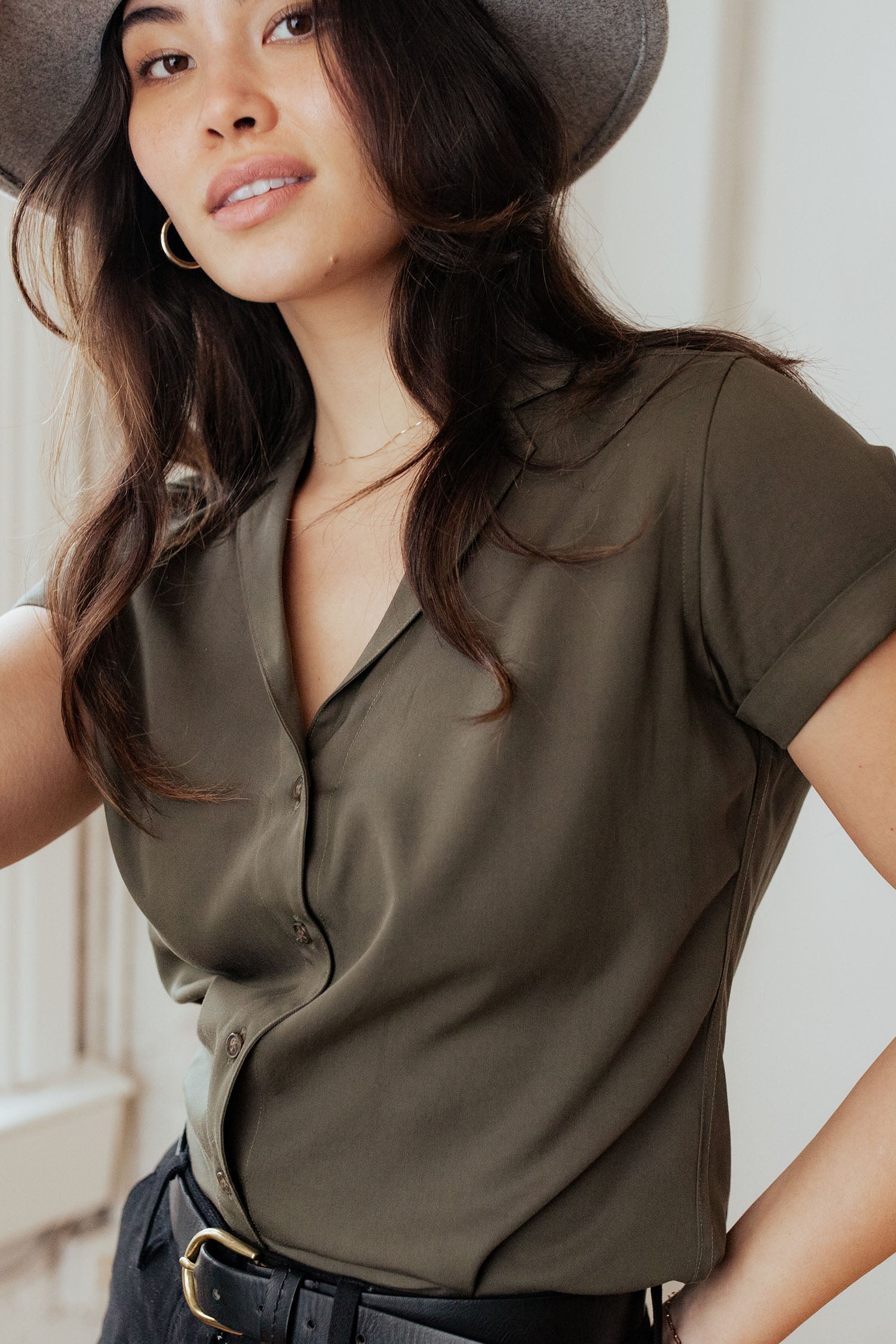 A woman stands indoors, looking at the camera with a slight smile, wearing the Bridge & Burn Innes Shirt in Olive, paired with a black belt and a gray wide-brim hat.