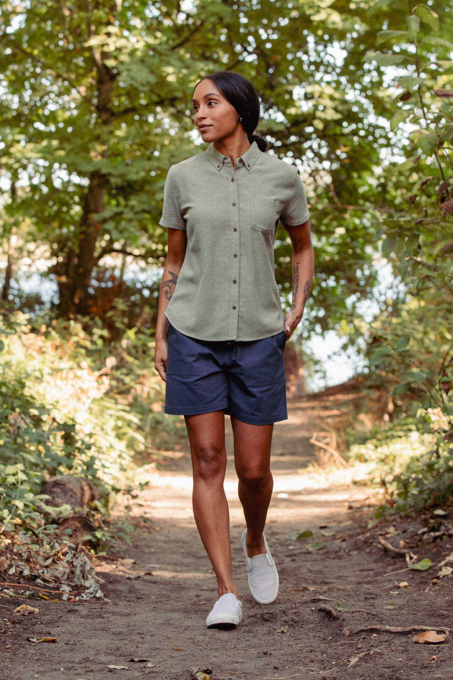 Wearing the Bridge & Burn Juniper Shirt in Olive Twill, a woman strolls a forest trail in navy shorts and white slip-ons, her relaxed fit blending effortlessly with the lush greenery.