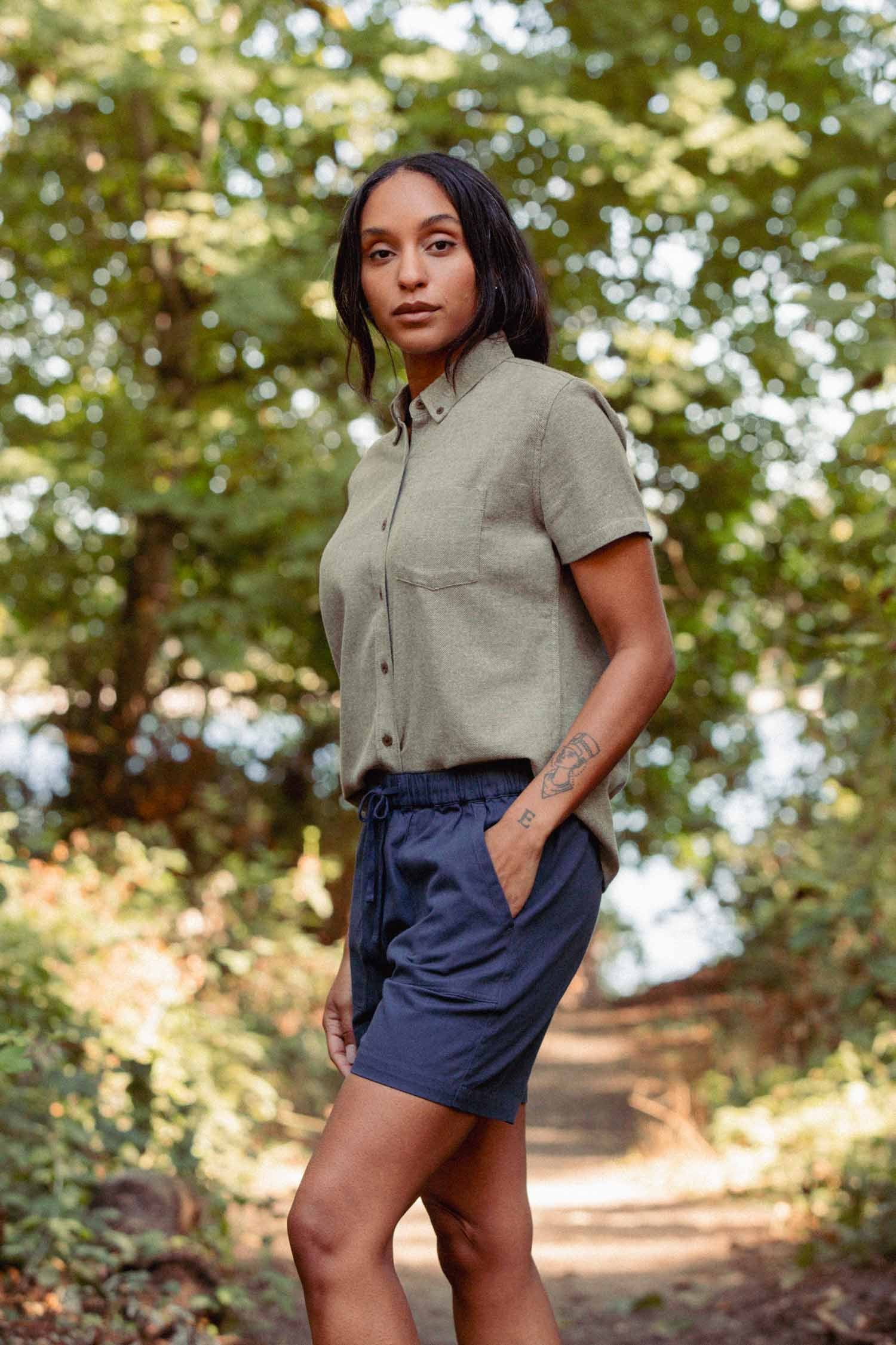 A woman stands on a wooded path wearing the Bridge & Burn Juniper Shirt in Olive Twill and navy shorts. Sunlight filters through the trees as she calmly looks at the camera, one hand in her pocket.