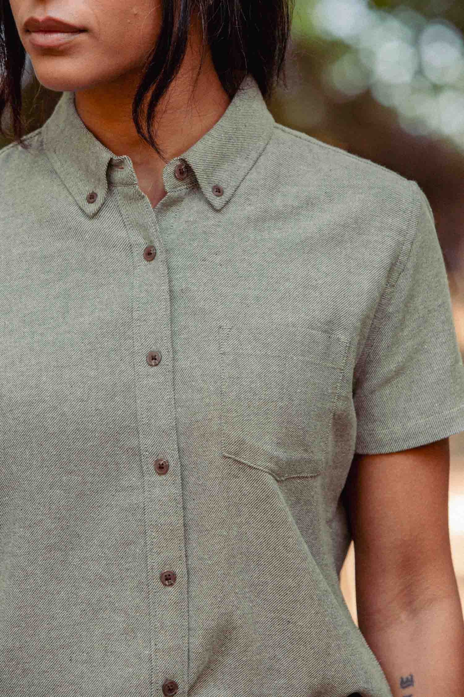 A person wears the Bridge & Burn Juniper Shirt in Olive Twill—a relaxed fit, short sleeve button-down with a chest pocket, crafted from recycled cotton—shown from shoulders to waist against a blurred green and brown background.