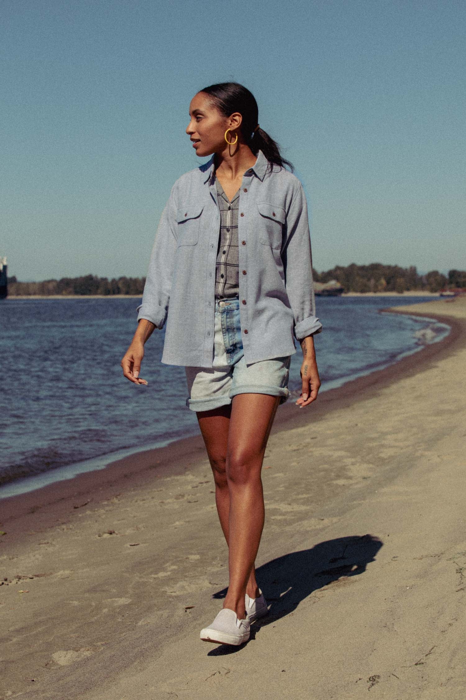 A woman walks by the water on a sandy beach, wearing the Bridge & Burn Lenon Shirt in denim twill over a striped top, with denim shorts and white slip-on shoes. Large hoop earrings complete her look, with trees and blue sky behind her.