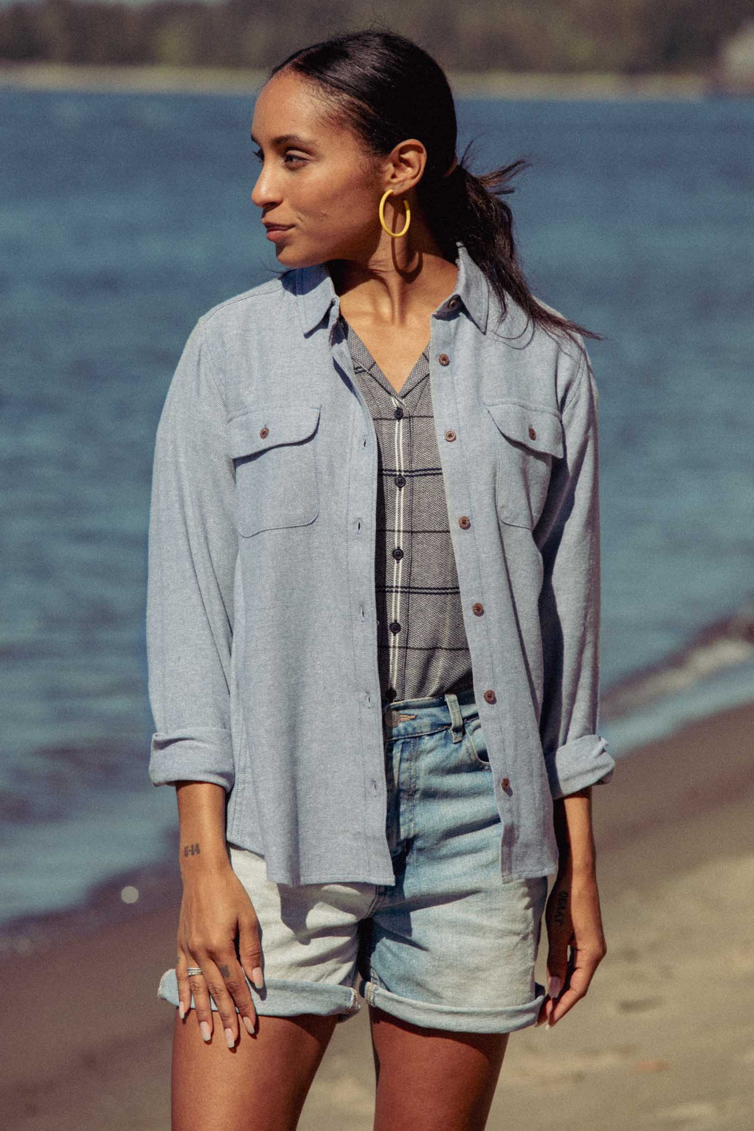 A woman stands on a sandy beach near the water, wearing the Lenon Shirt in Denim Twill by Bridge & Burn over a plaid top and denim shorts, accessorized with large hoop earrings as she gazes to the side.