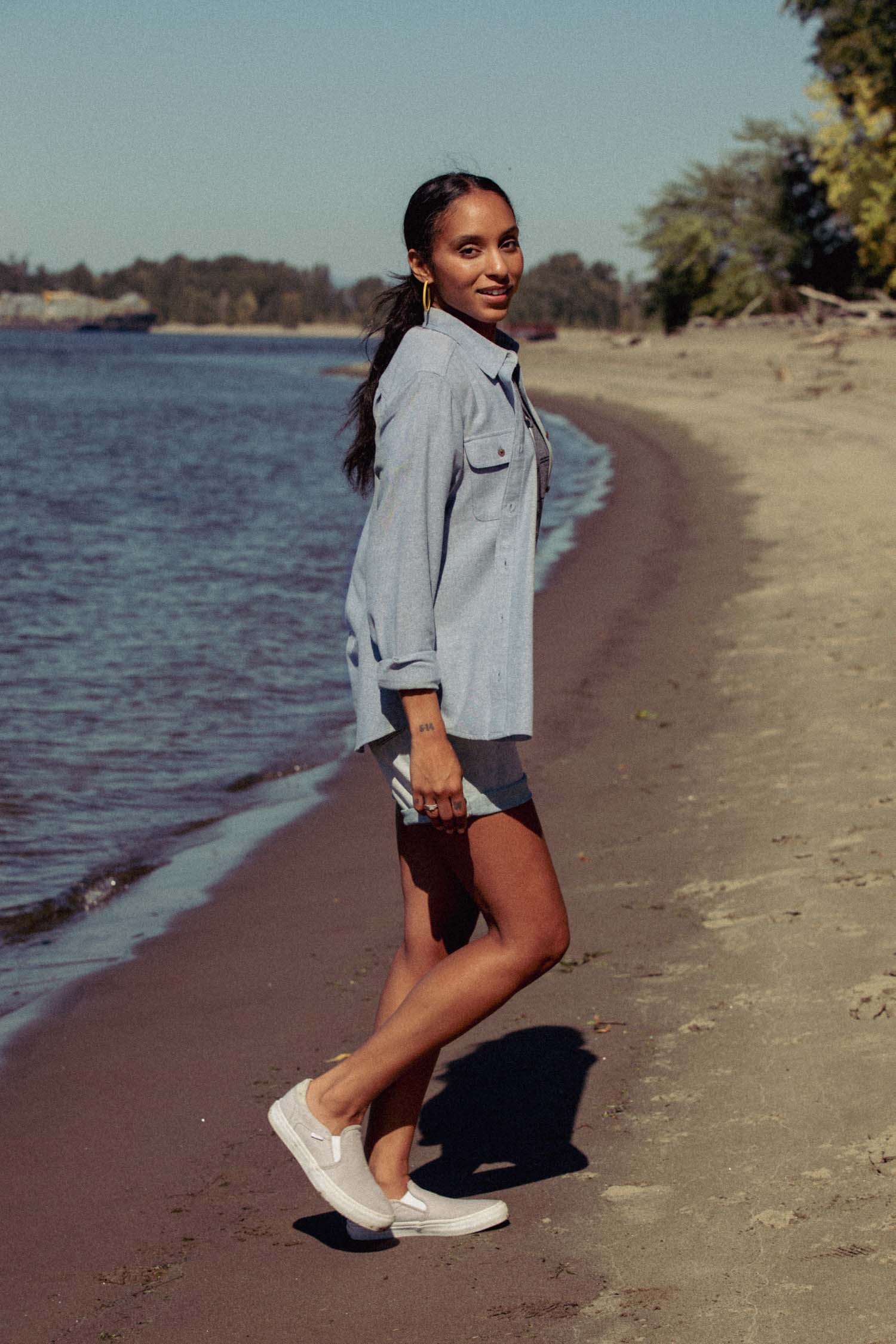 A woman walks barefoot along a sandy beach near the water, smiling as she looks back. She wears the Bridge & Burn Lenon Shirt in Denim Twill and shorts, with trees and a clear sky in the background.