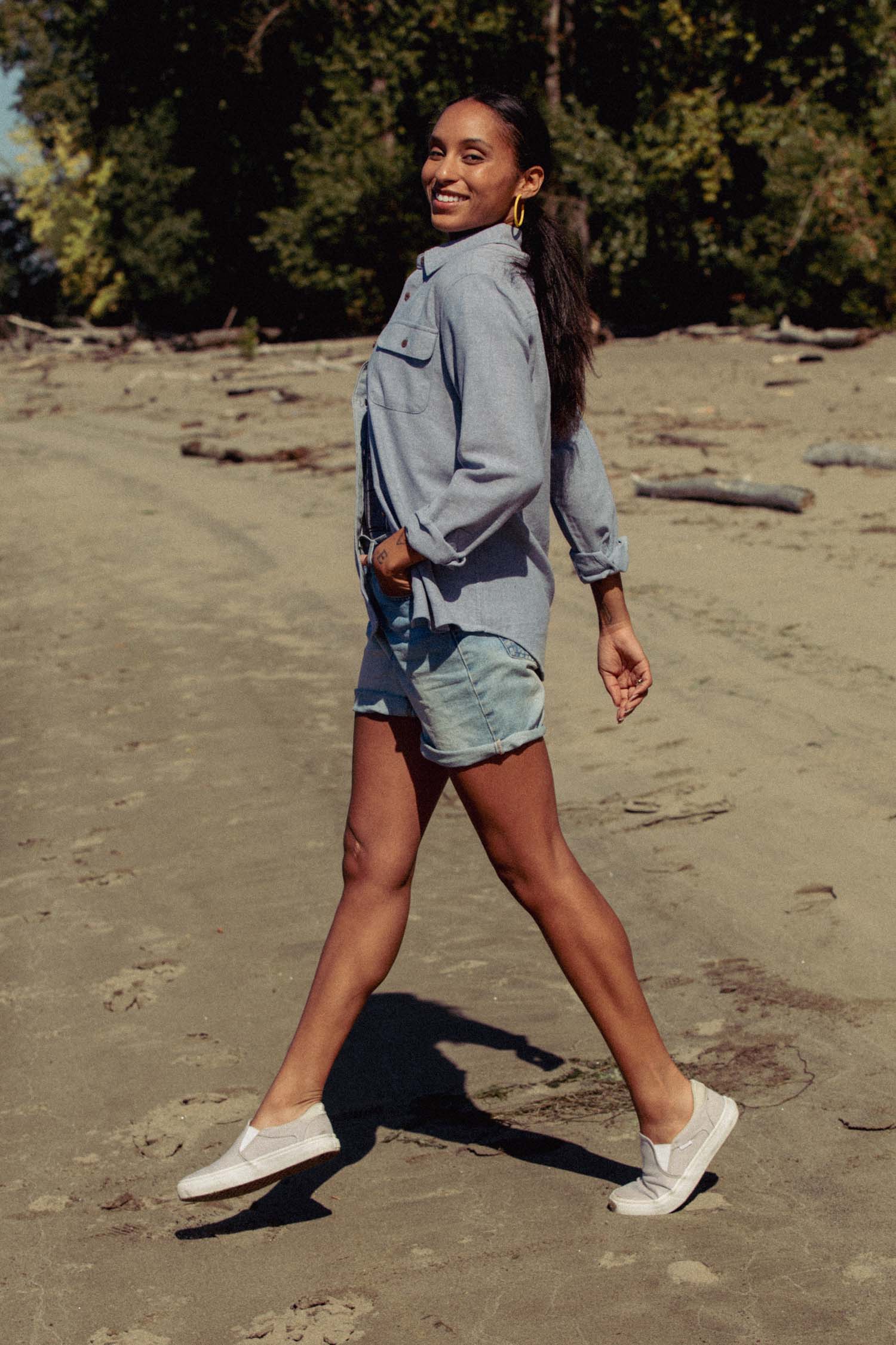 A woman wearing the Bridge & Burn Lenon Shirt in Denim Twill strolls and smiles on a sandy beach, with trees in the background.