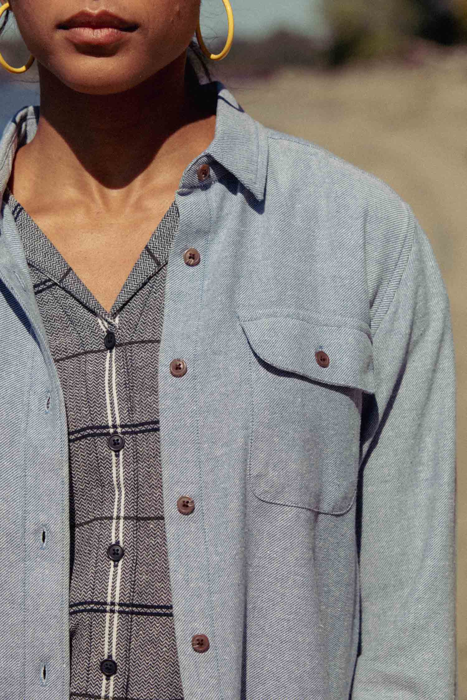 Wearing the Bridge & Burn Lenon Shirt in Denim Twill layered over a gray, vertically striped dress, this person accessorizes with gold hoop earrings; only the lower half of their face is shown.